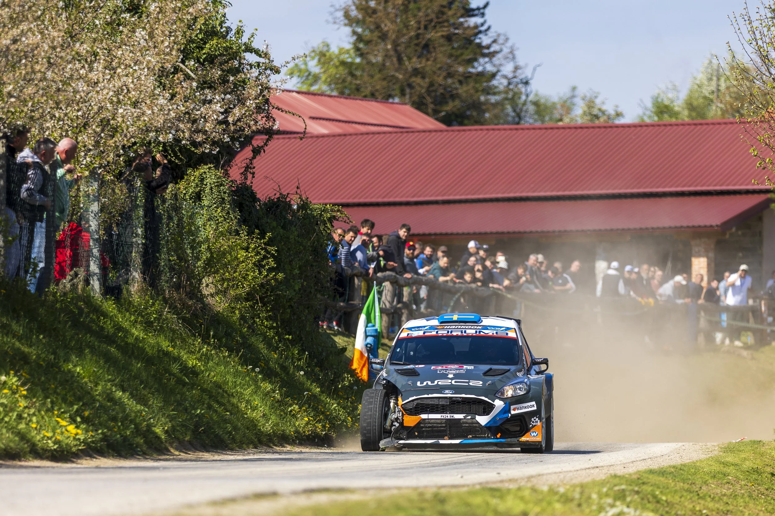 A rally car racing on a dirt road with spectators watching from the side, some standing behind a fence and others on a grassy bank. The car is kicking up dust as it speeds past, with a red-roof building and trees in the background, during the daytime