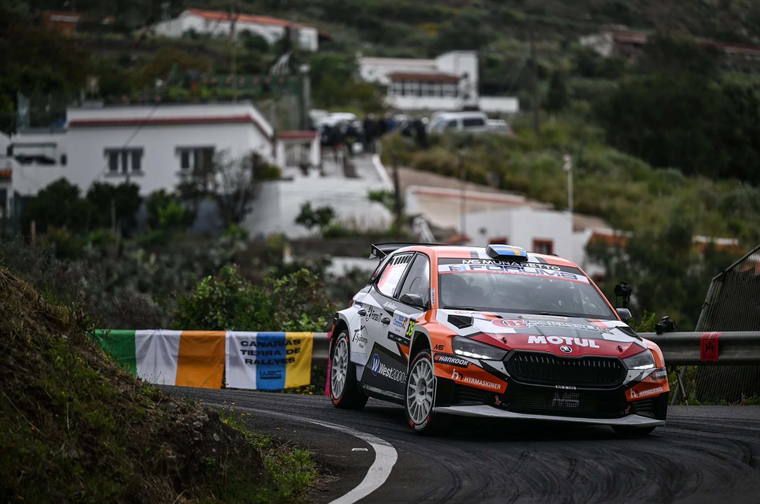 Rally car on a winding mountain road during a race, with houses and lush green hills in the background.