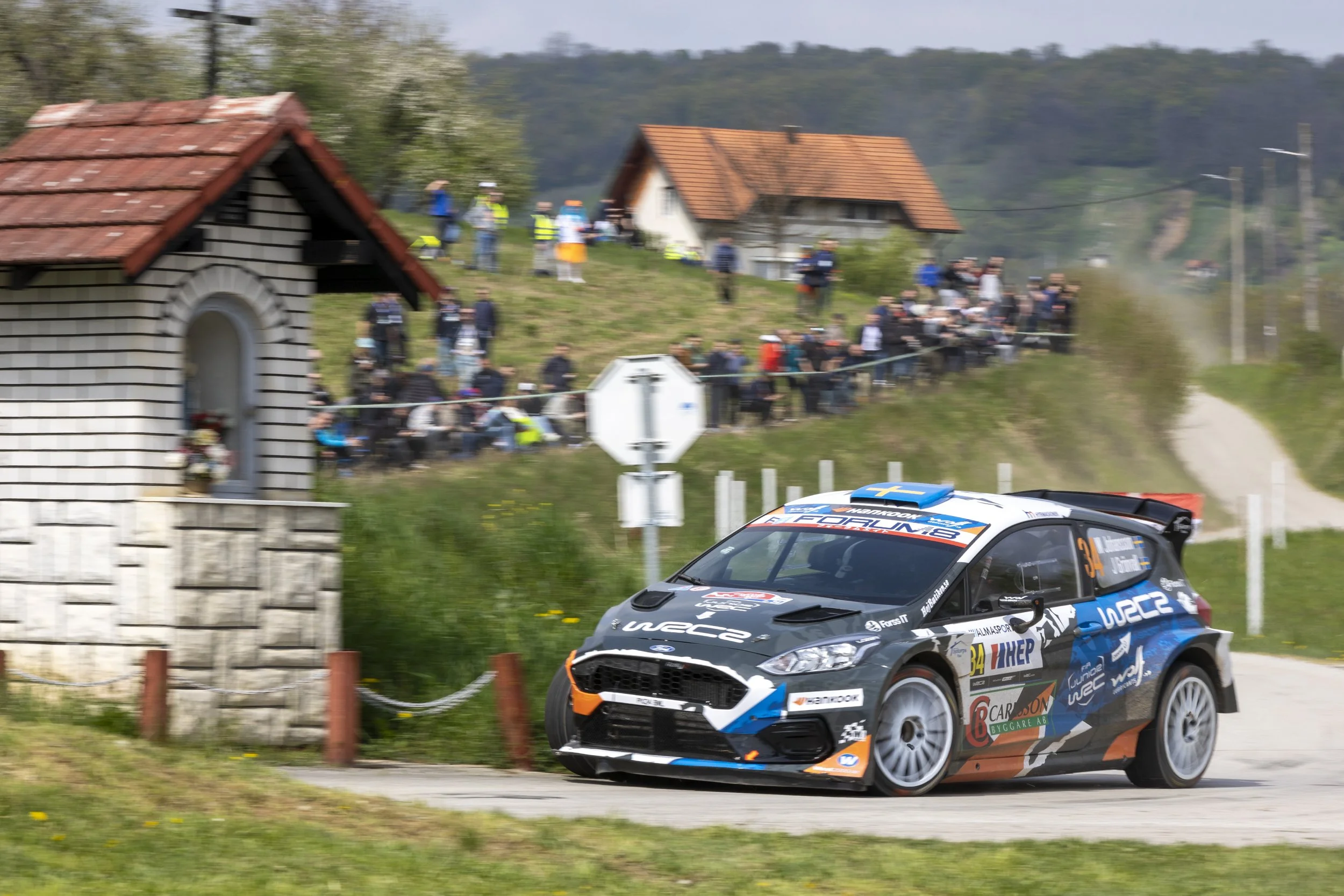A rally car racing on a dirt road with a crowd of spectators standing on a grassy hill in the background, and a small brick building with a red tile roof nearby.