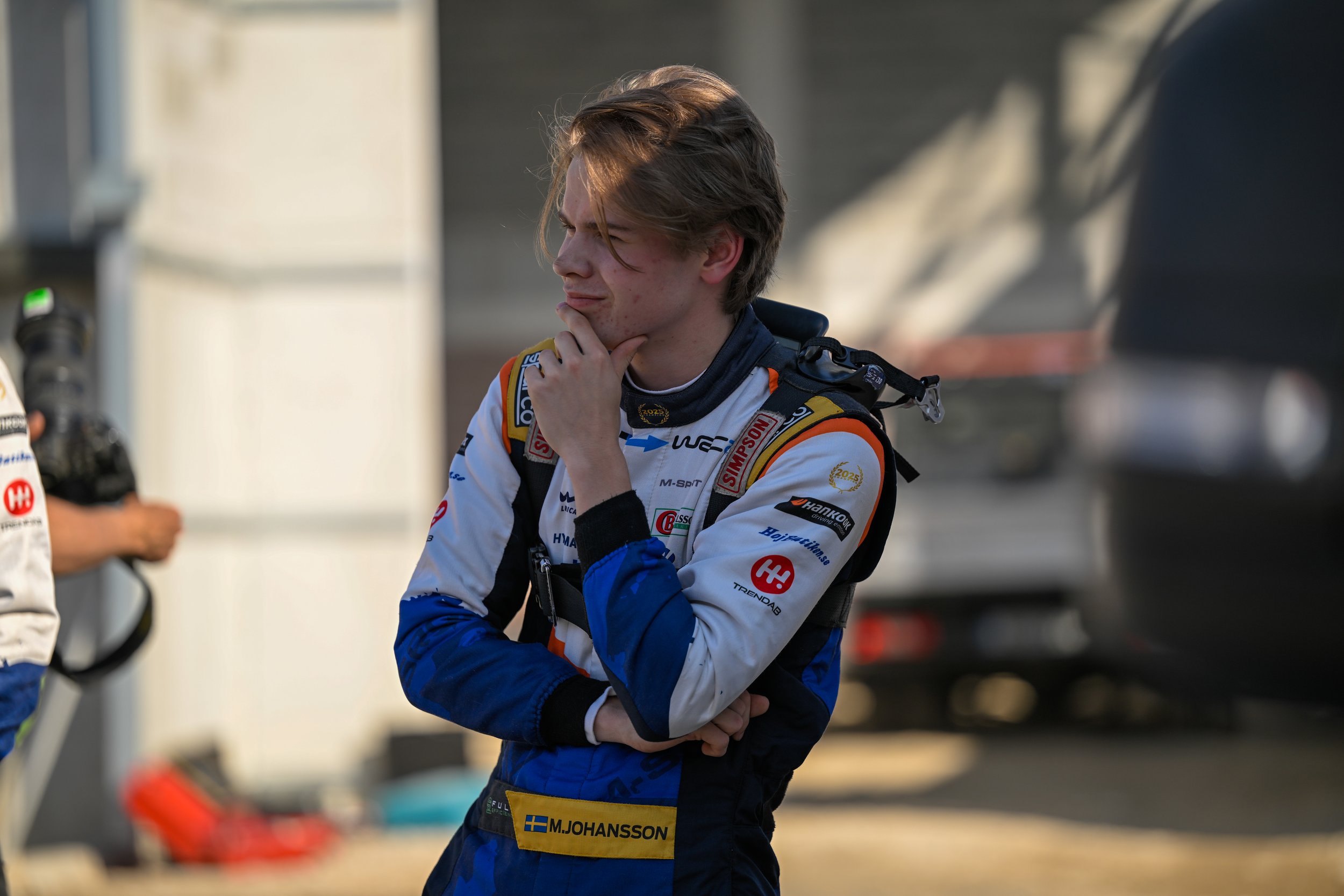 Young male race car driver in racing suit, standing outdoors, looking thoughtful with his hand on his chin.