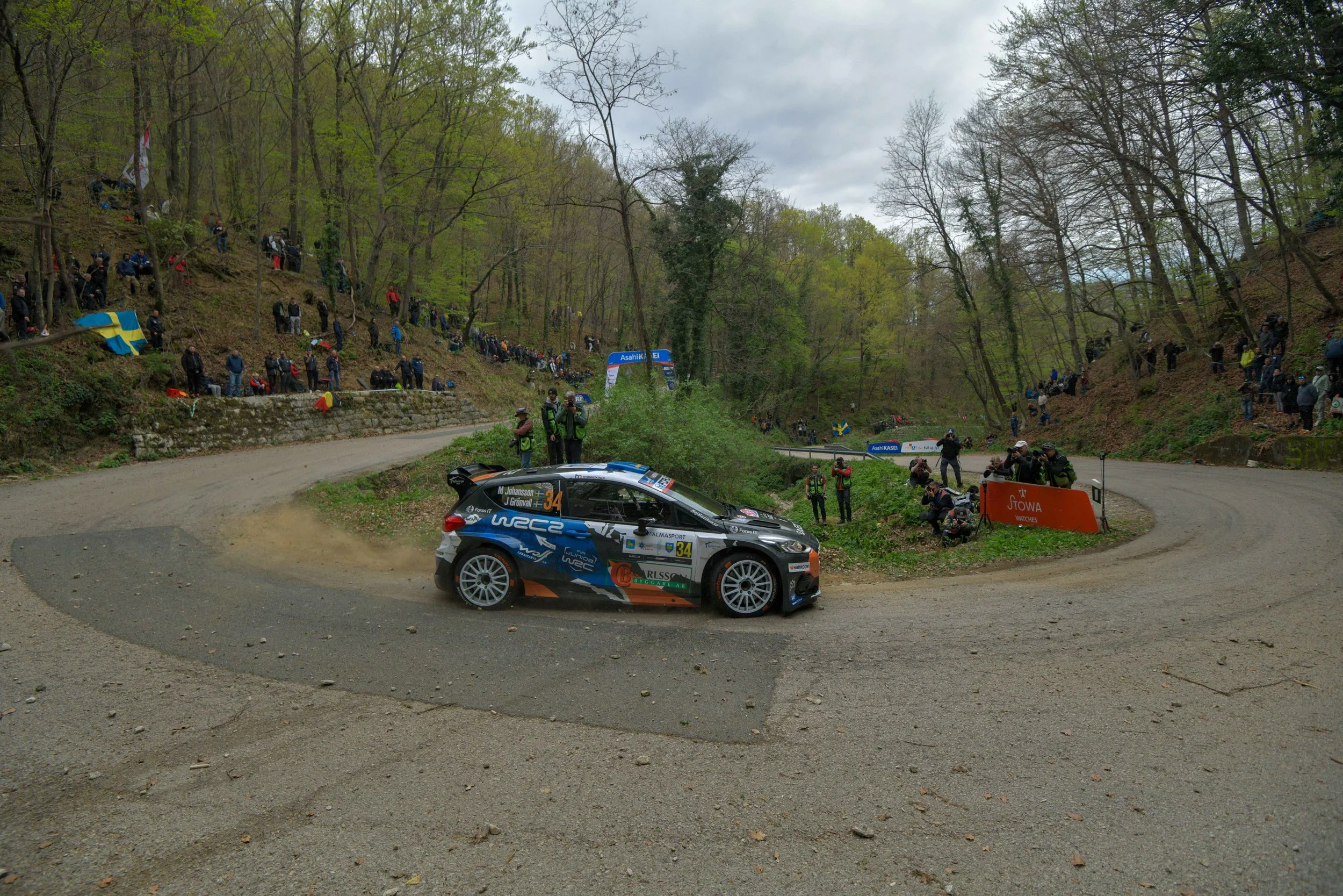 A rally car speeding on a dirt road during a race, with spectators lined up along the hillside and trees in the background.
