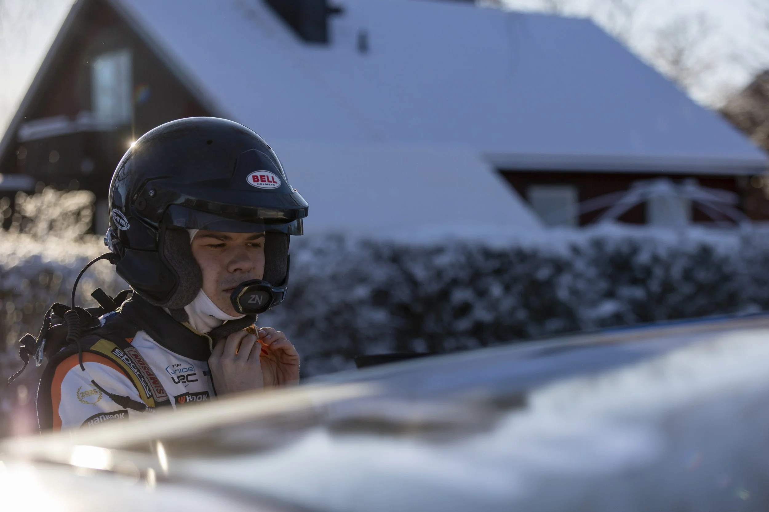 A race car driver in a racing suit and helmet preparing for a race, with a snowy landscape and a red barn in the background.
