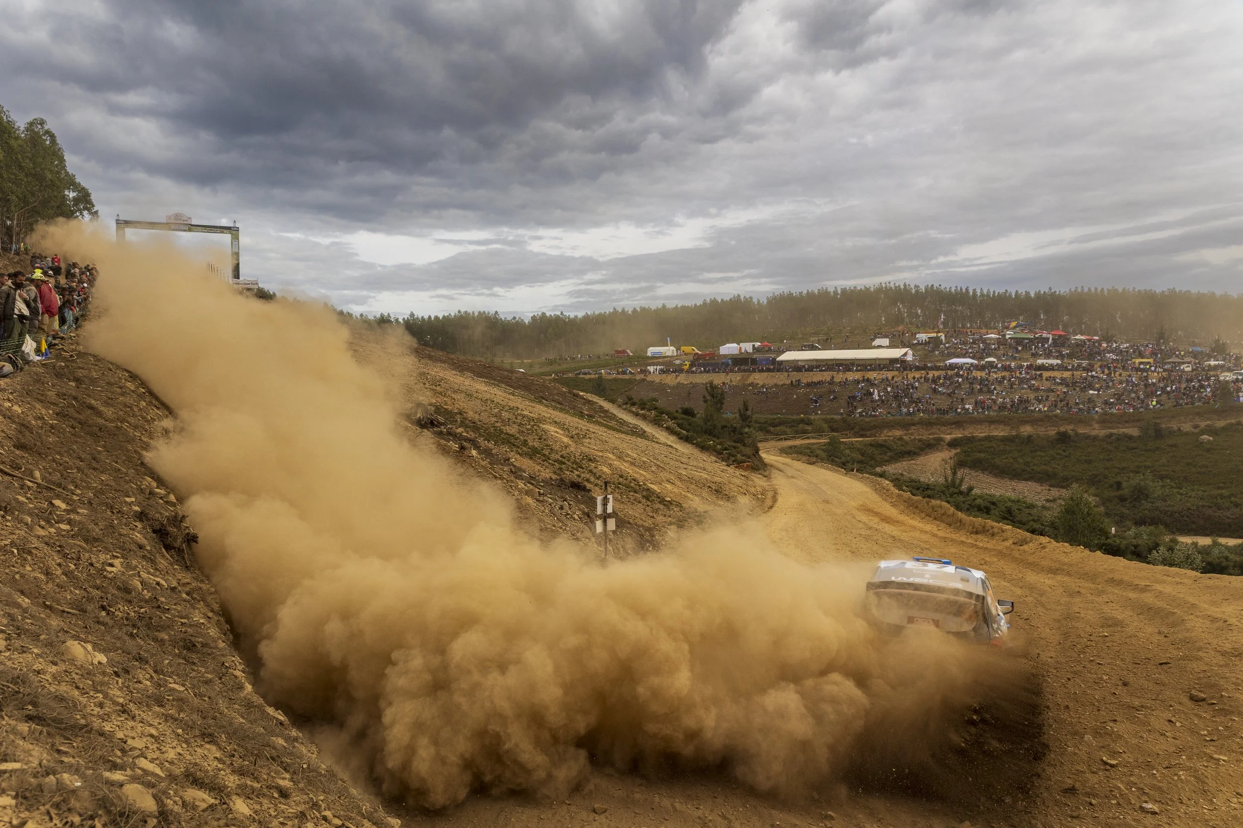 Race cars racing on a dirt track during a rally event with a large crowd and tents in the background, cloudy sky overhead.