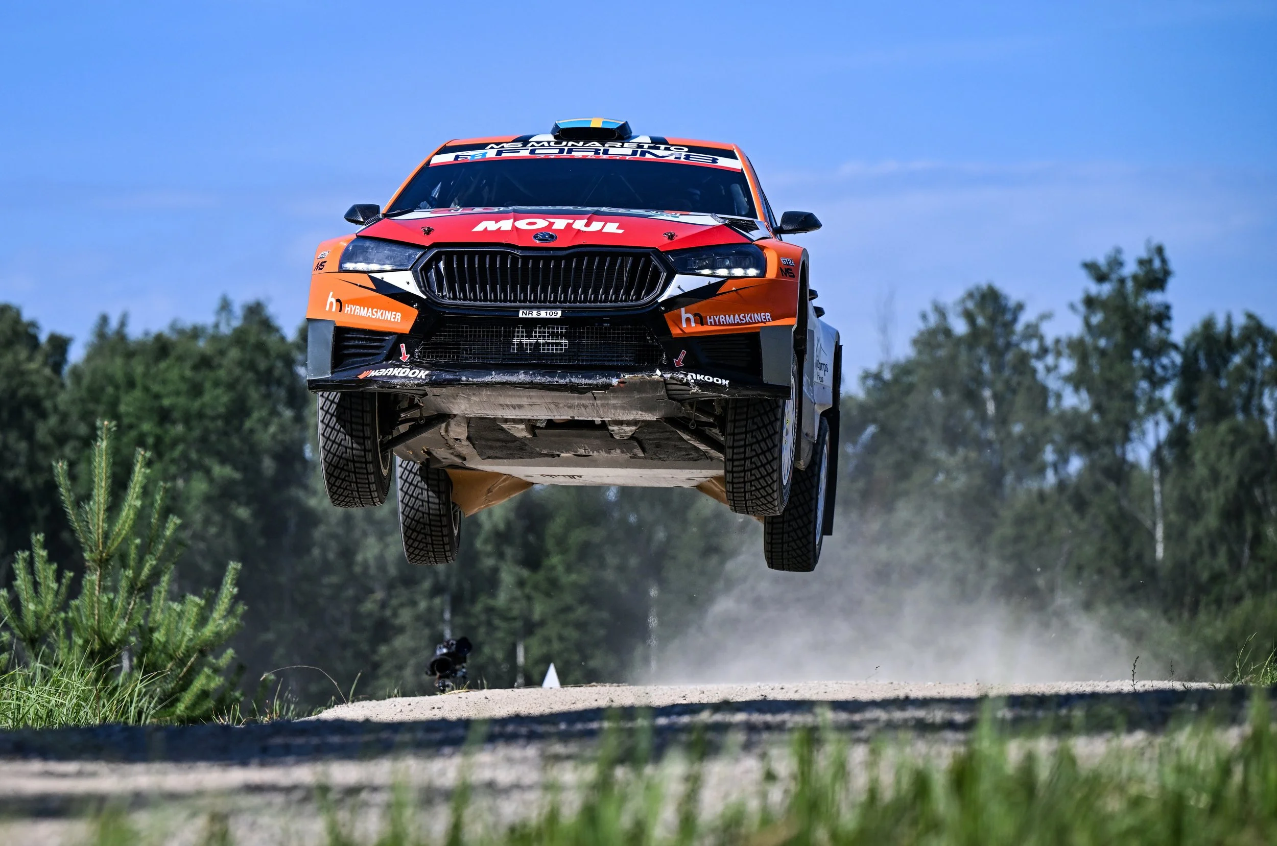 A rally car flying in the air above a dirt road during a race, with a blue sky and trees in the background.