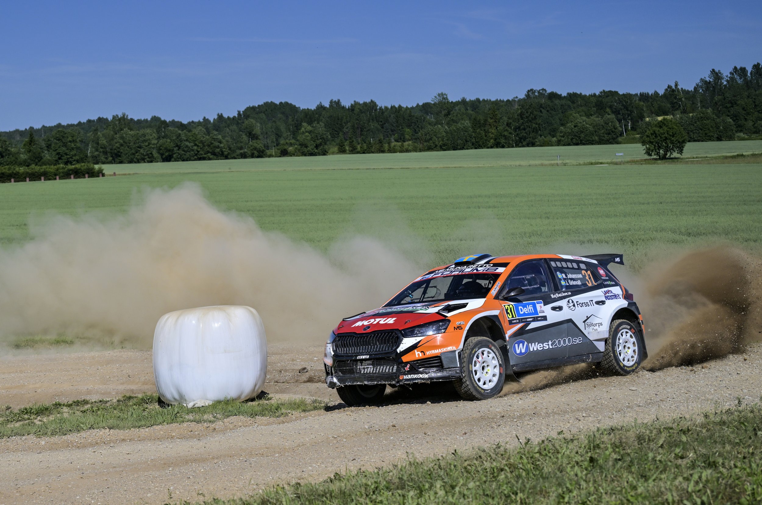 A rally car racing on a dirt track, kicking up dust behind a white hay bale, with lush green fields and trees in the background under a clear blue sky.