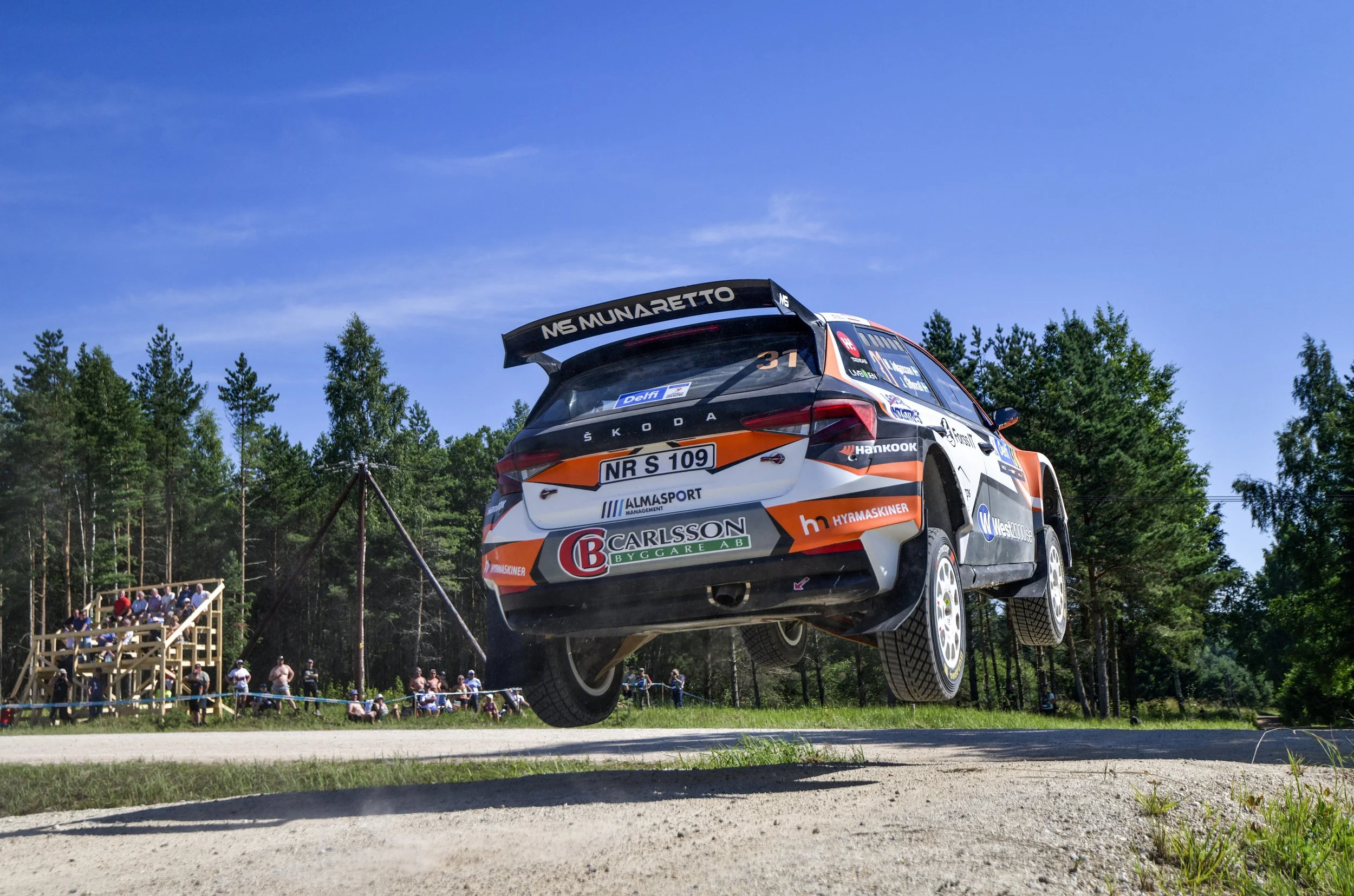 A rally car is airborne during a jump at a motorsport event, with spectators watching from a wooden stand and a forested area in the background.