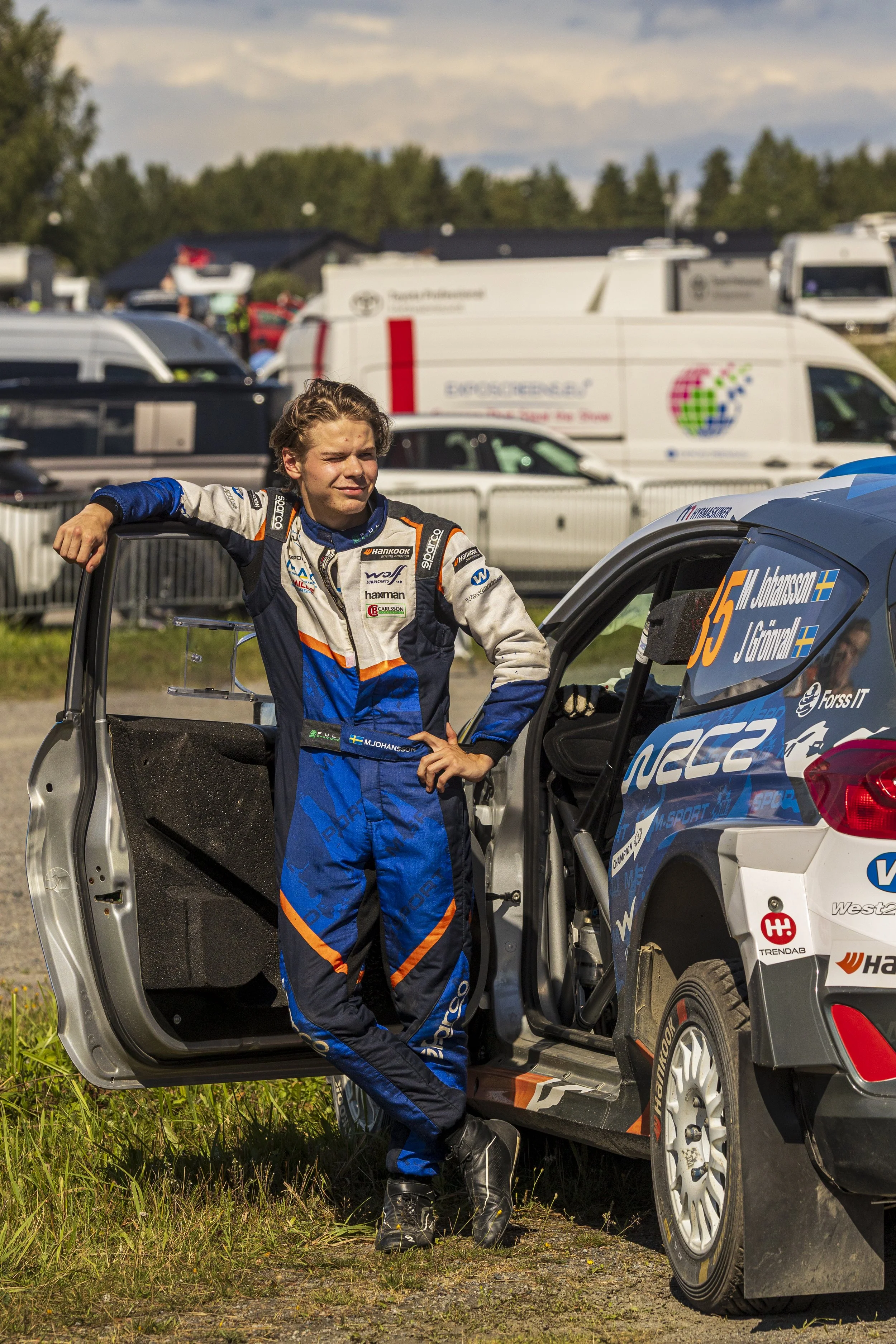 A young male rally car driver in a racing suit standing with one hand on his hip, next to a rally car with the door open, in an outdoor setting with grass and a parking lot full of vehicles in the background.