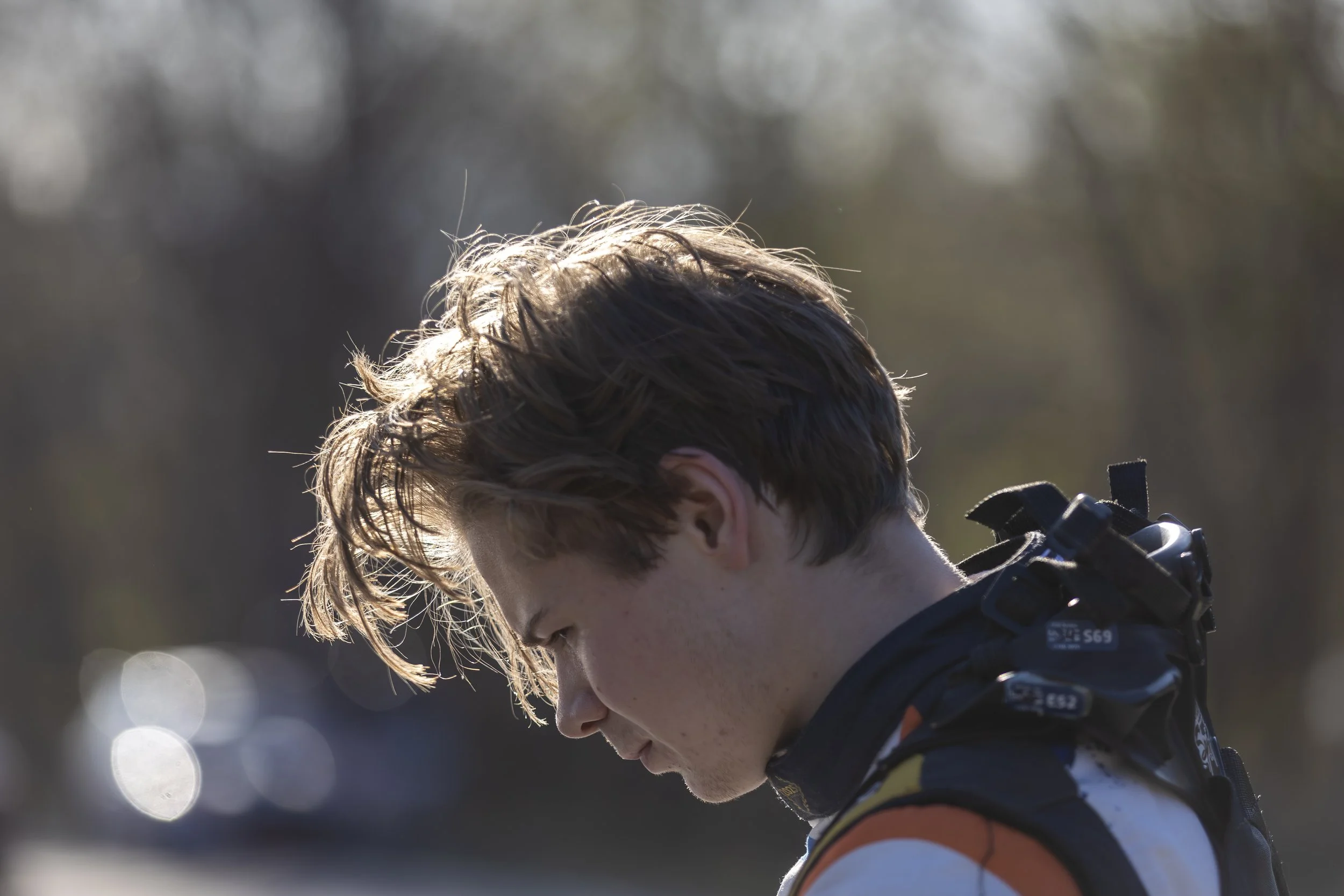 Side profile of a young man with light brown hair wearing a black sports helmet and orange and white sports attire, standing outdoors in sunlight with a blurred background.