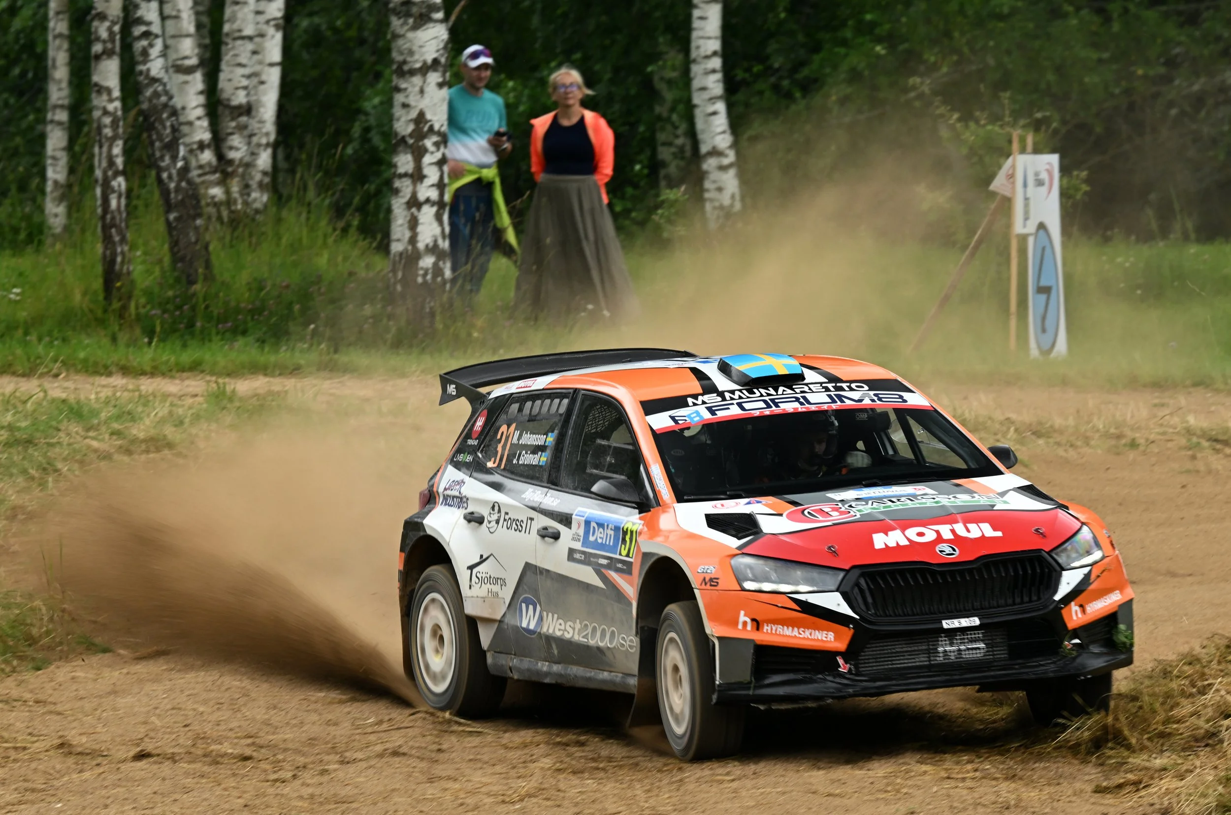 A rally car speeding on a dirt road in a wooded area, kicking up dust, with two spectators standing behind trees watching the race.
