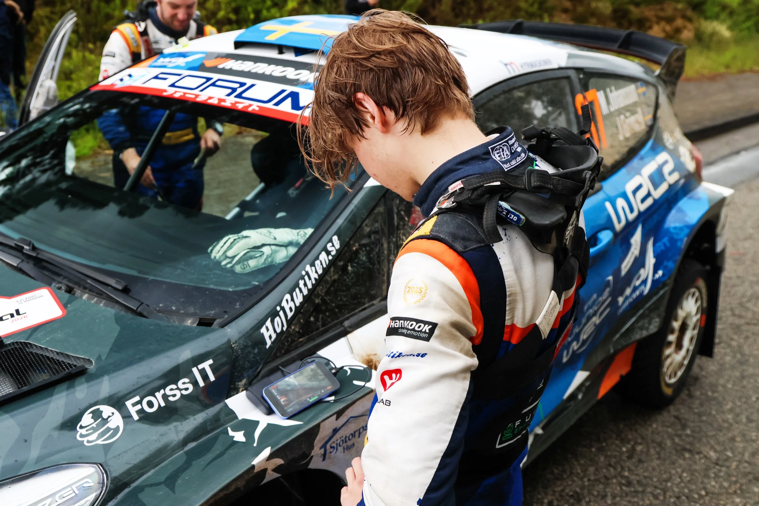 A rally car with racing decals and a driver and co-driver preparing next to it on a dirt road.