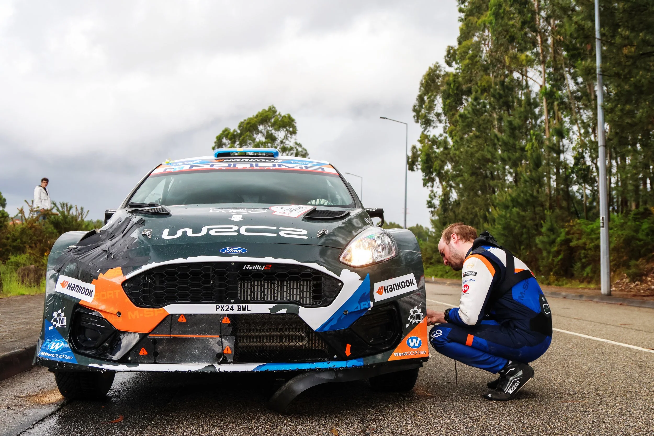 A black rally car with sponsor logos, a person in a racing suit crouching and working on the car's front, and a person in casual clothes standing in the background on a cloudy day with trees.