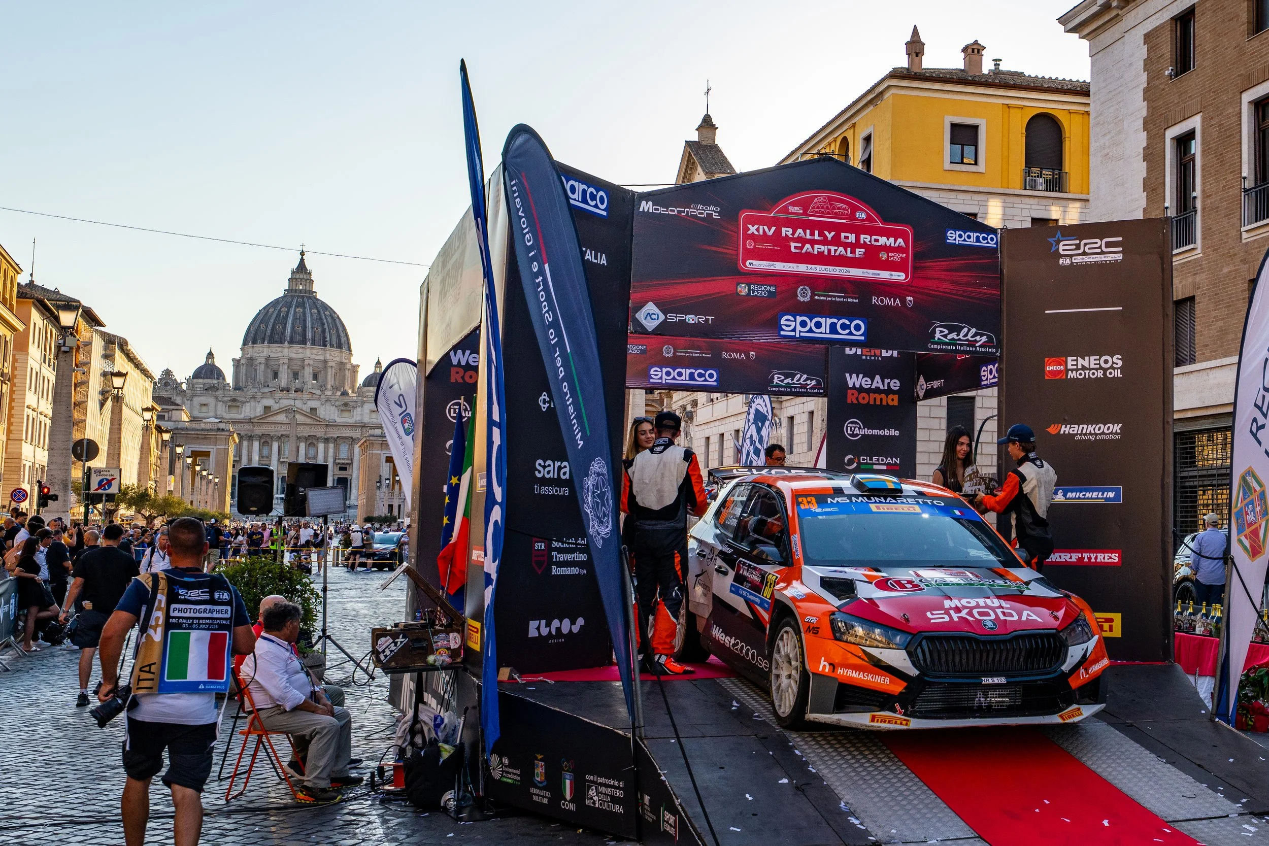 A rally car on a platform at a racing event in an urban square with historic buildings and a domed church in the background. The scene includes banners, flags, and people, with some crew members inspecting the car and spectators watching.