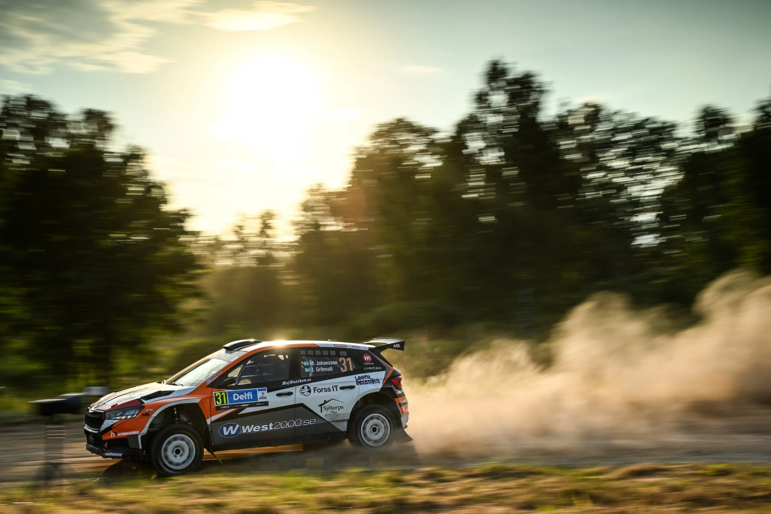 A rally car racing on a gravel road at high speed with dust trailing behind, with a blurred green forest and blue sky in the background.