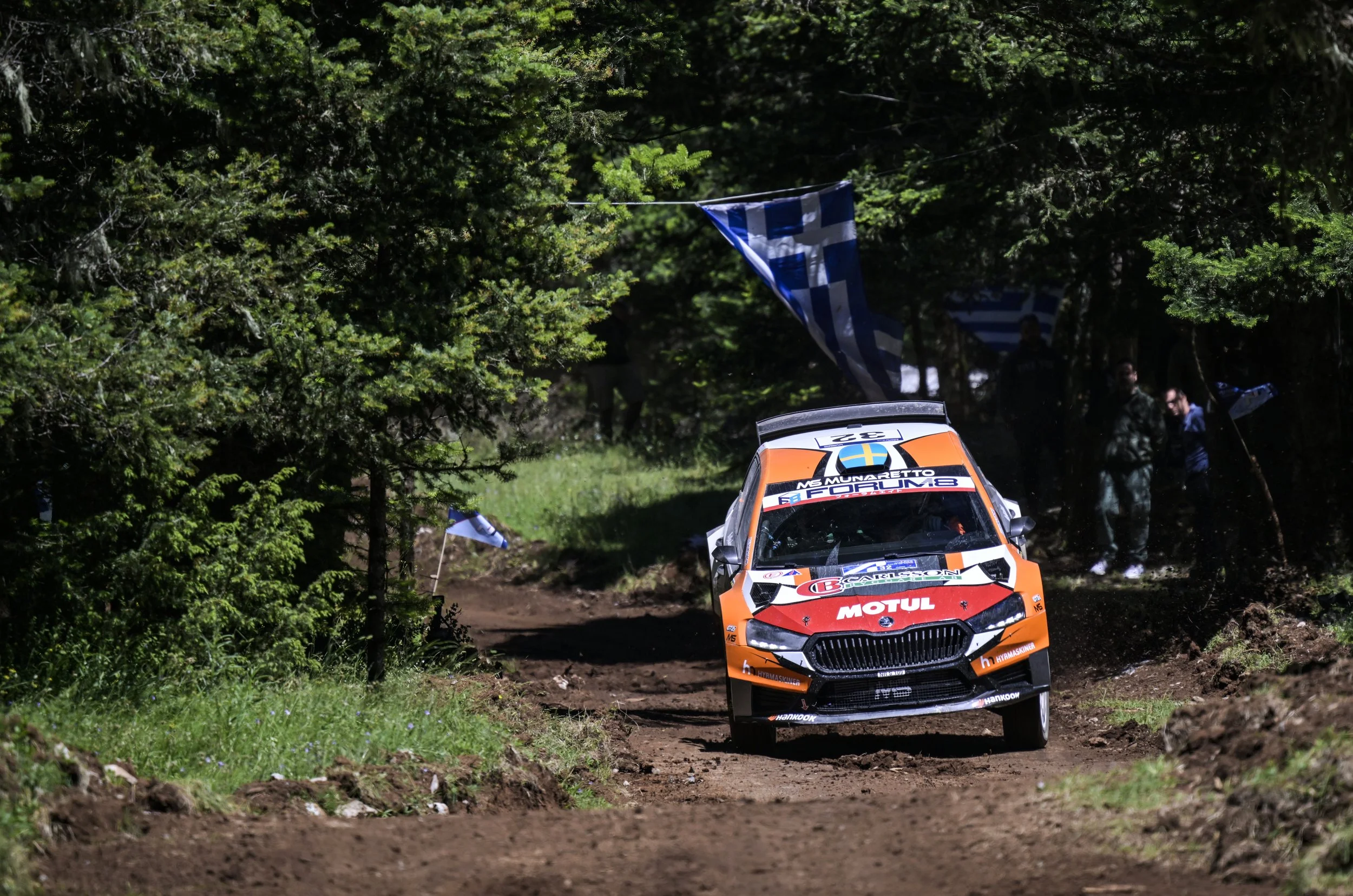 Rally car driving on a dirt forest trail with a Greek flag hanging from the trees and spectators in the background.
