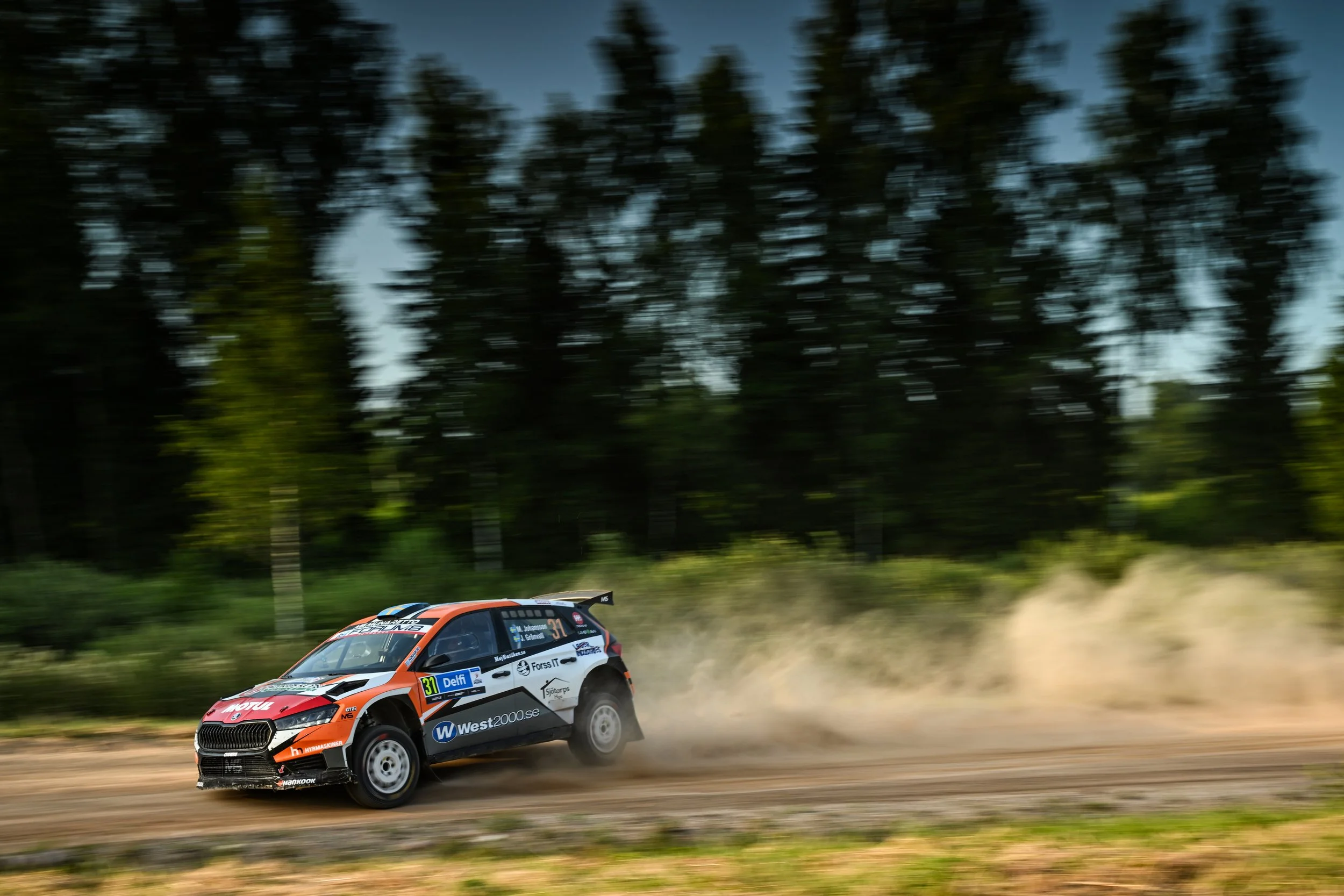 A rally car racing on a dirt track with a blurred background of trees, kicking up dust as it speeds past.