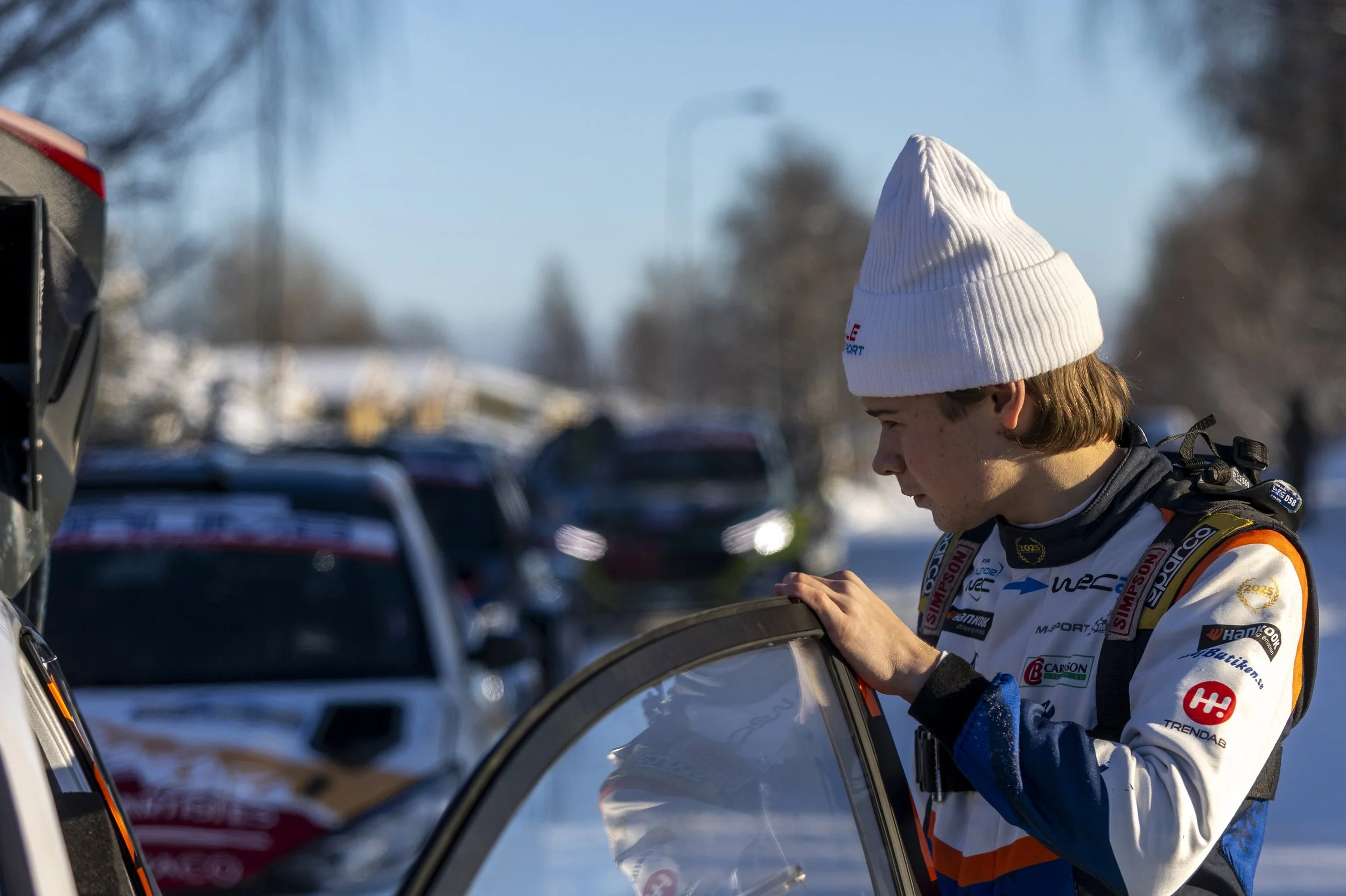 A person wearing a white beanie and a racing suit looking at a car door in a parking lot during winter.