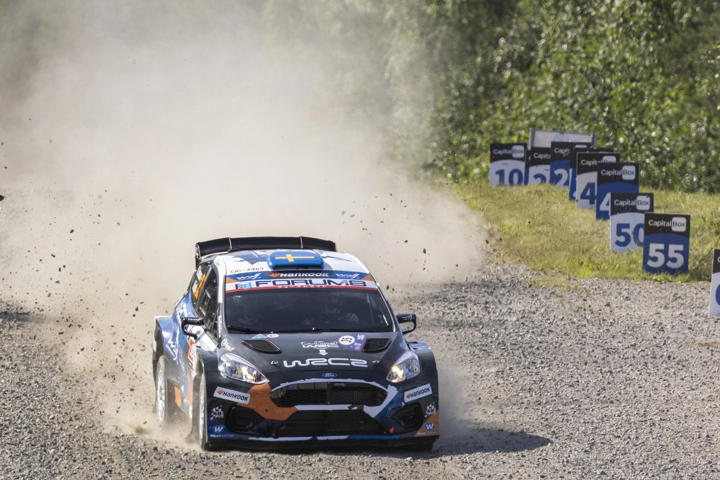 A rally car racing on a dirt track, kicking up dust and small rocks, with colorful advertising and racing numbers visible on the car and signs along the side of the track.