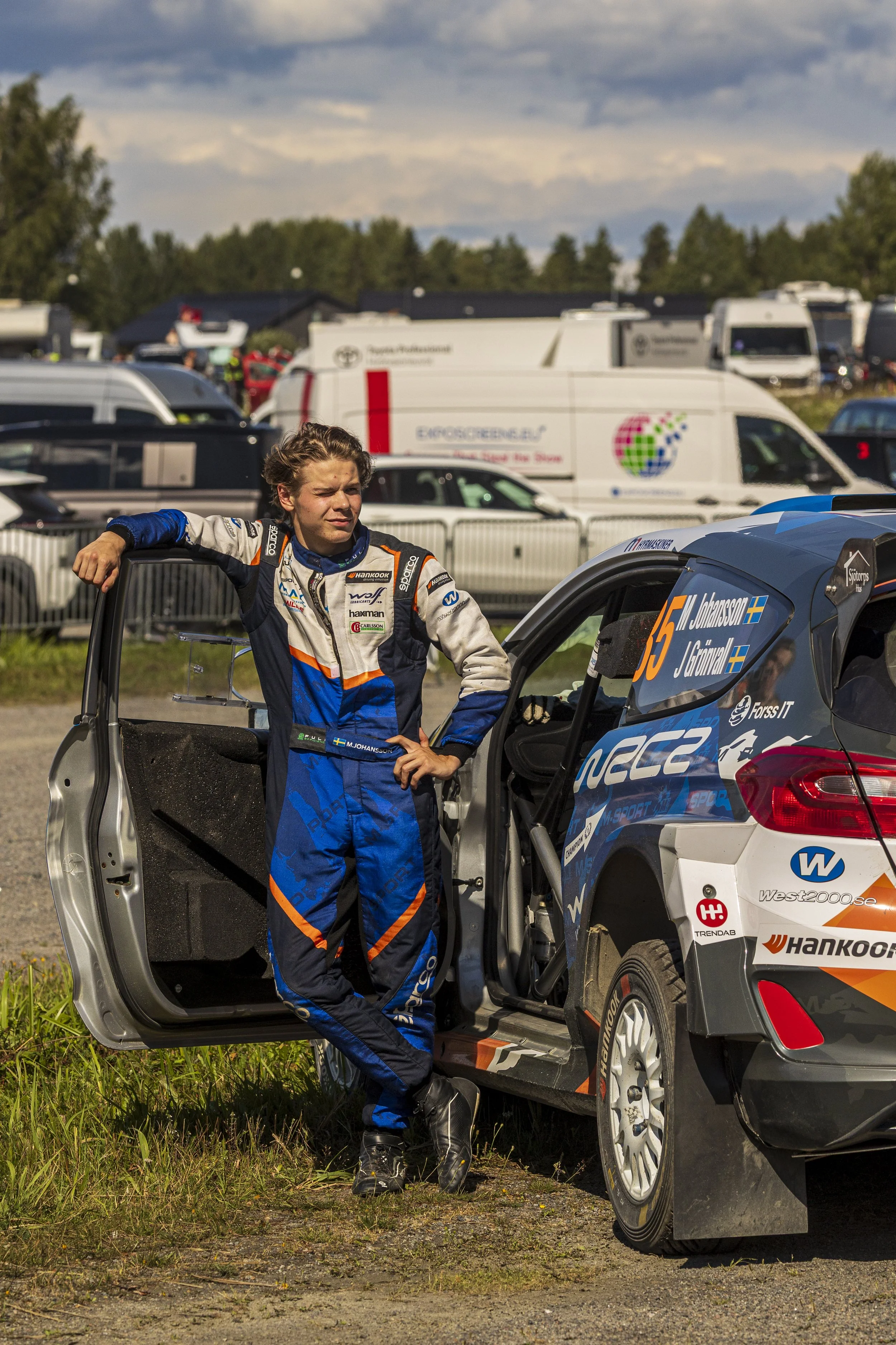 A young race car driver in a blue racing suit stands next to a race car with the door open at a racing event. The background shows various trailers and vehicles, with the sky partly cloudy.