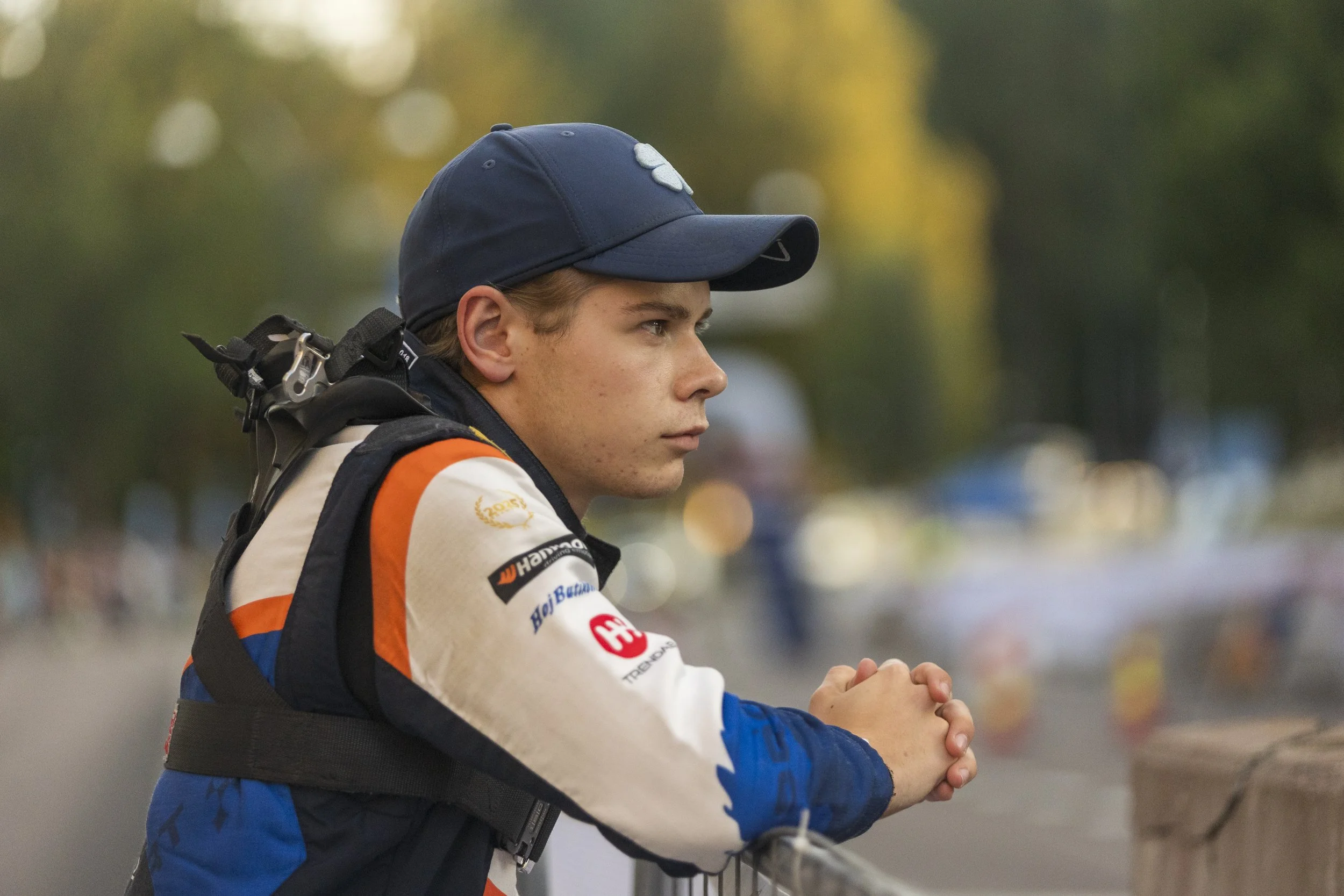 A young man with light brown hair wearing a navy blue cap, racing suit, and a racing harness, standing outdoors with his hands clasped together and looking to the right, with a blurred background of trees and lights.