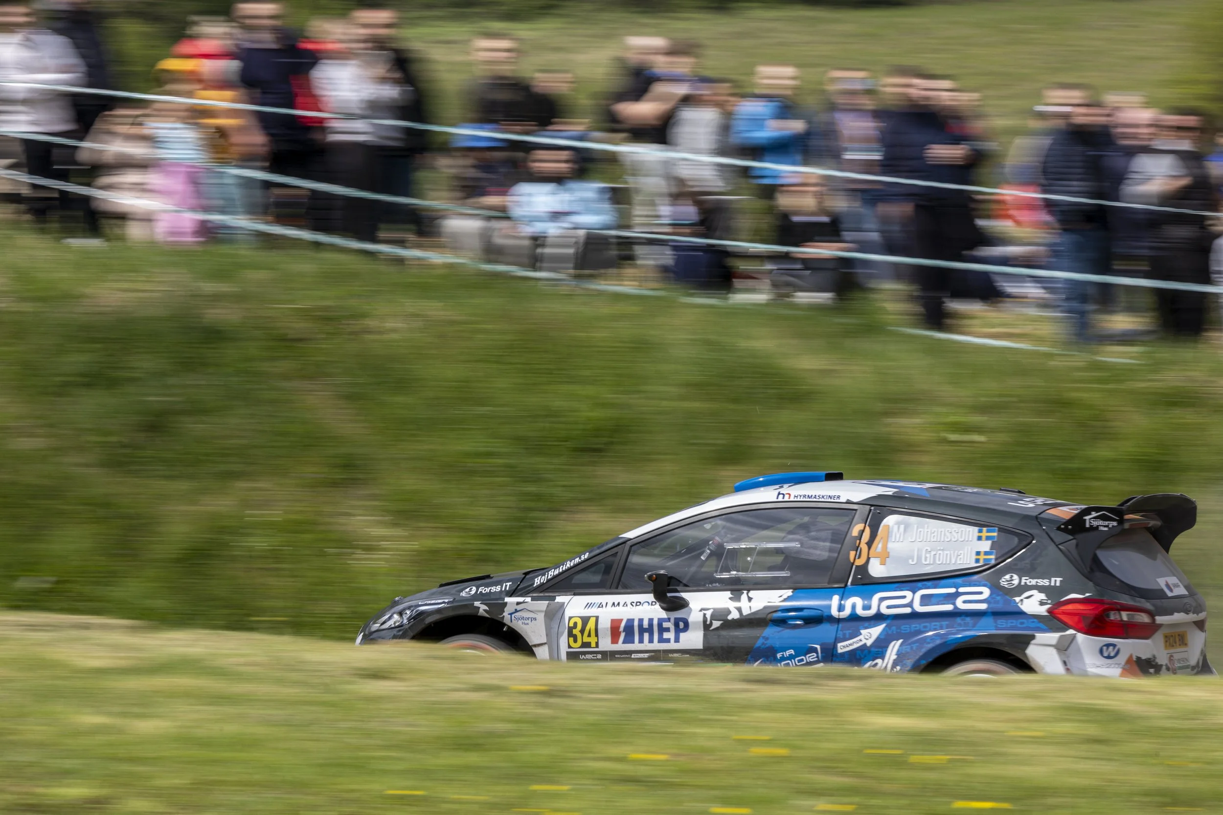 A rally car racing on a dirt track with a crowd of spectators blurred in the background.