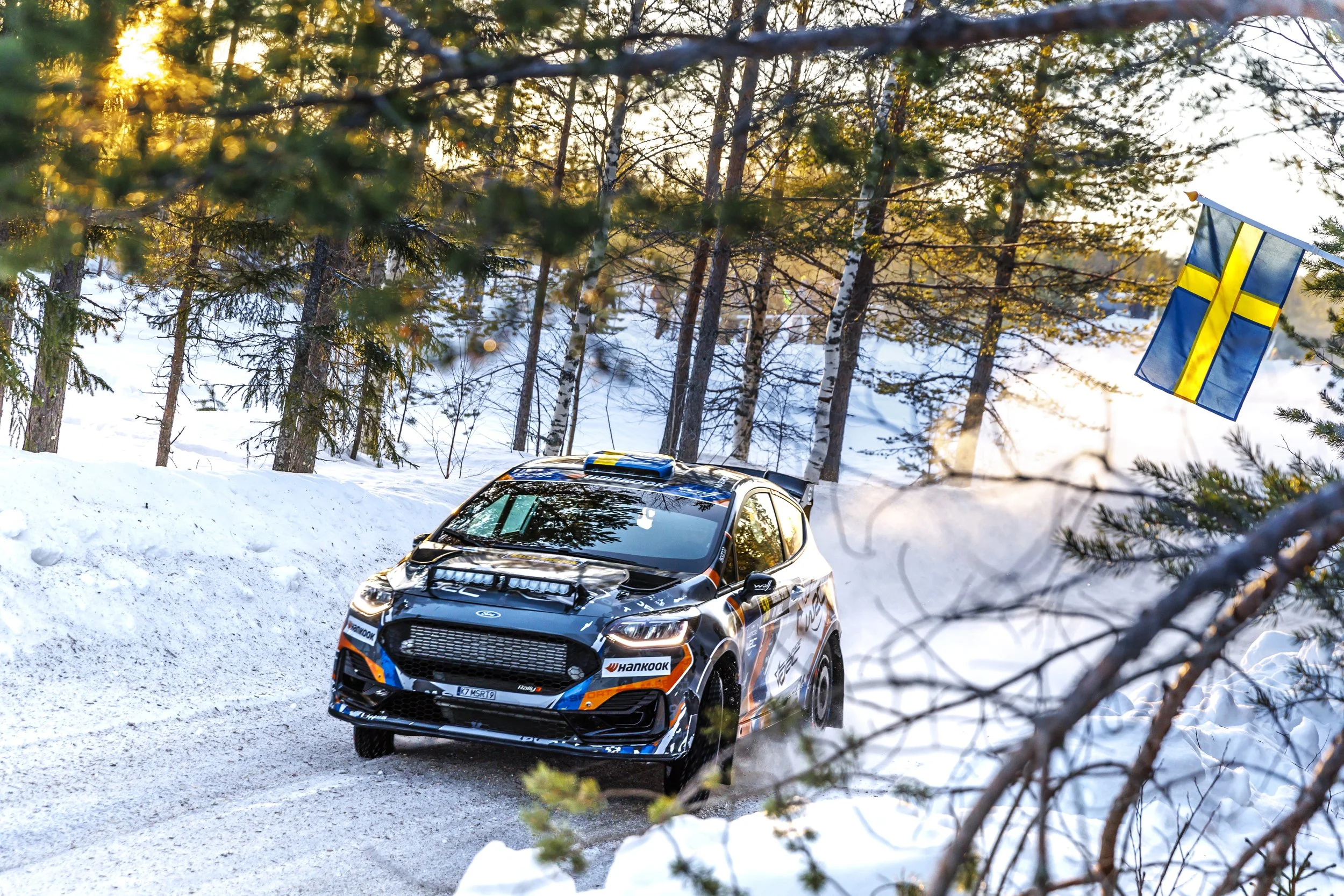 A rally car speeding on a snowy forest trail with a Swedish flag attached to a tree in the background, during sunset.