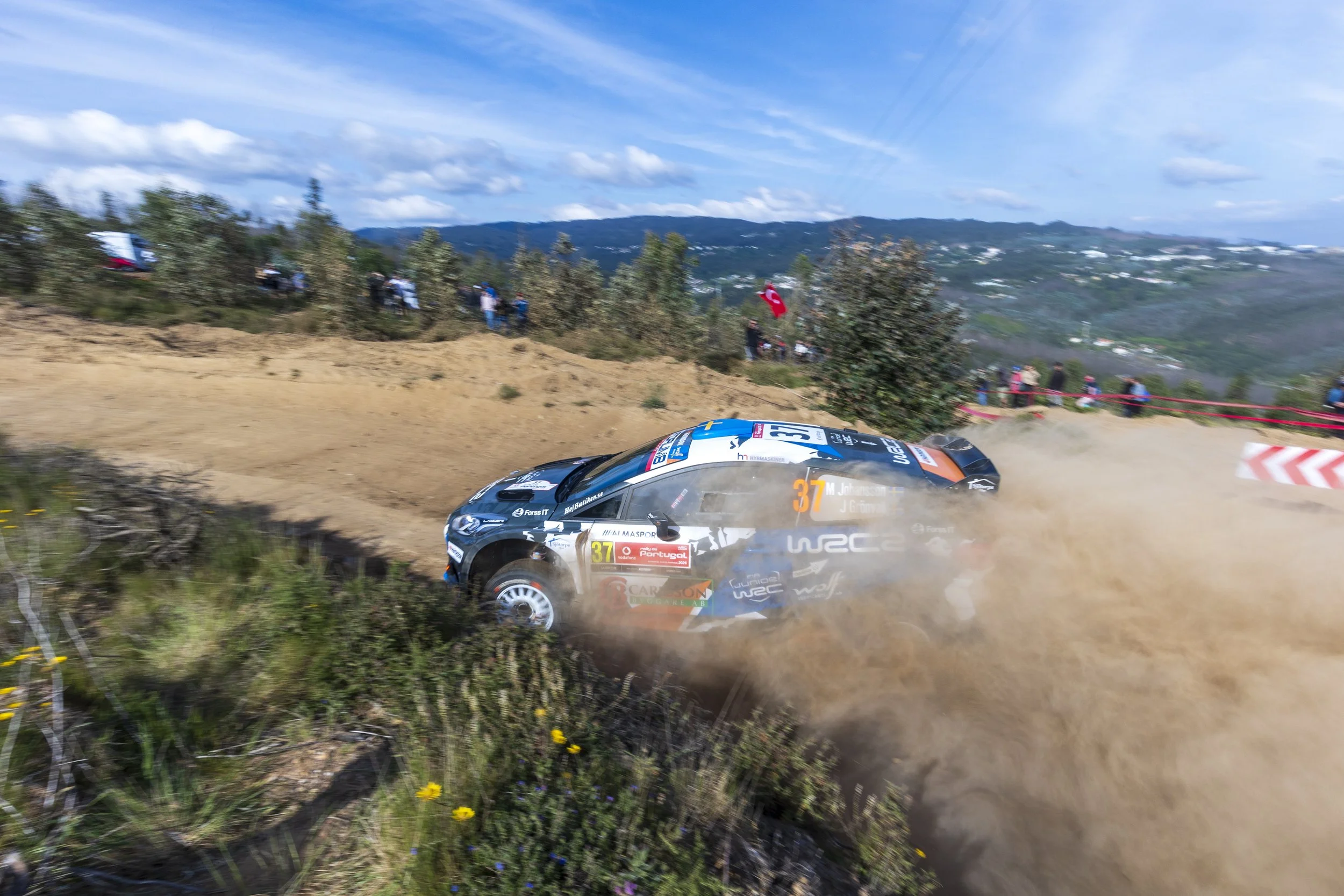 A rally car speeding on a dirt road with dust trailing behind, racing through a landscape with onlookers and a Turkish flag in the background.