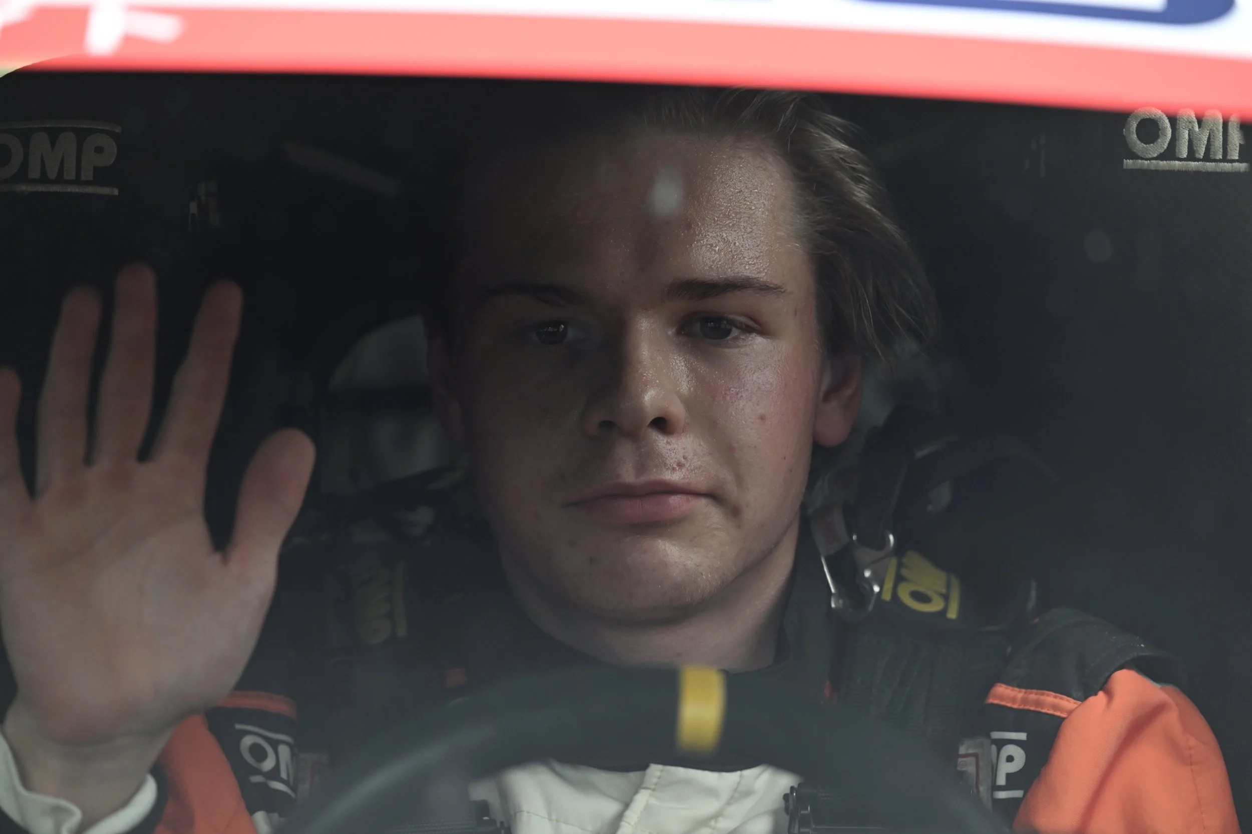 Race car driver with helmet visor up, wearing racing suit, inside racing car cockpit, hand raised in greeting or farewell.