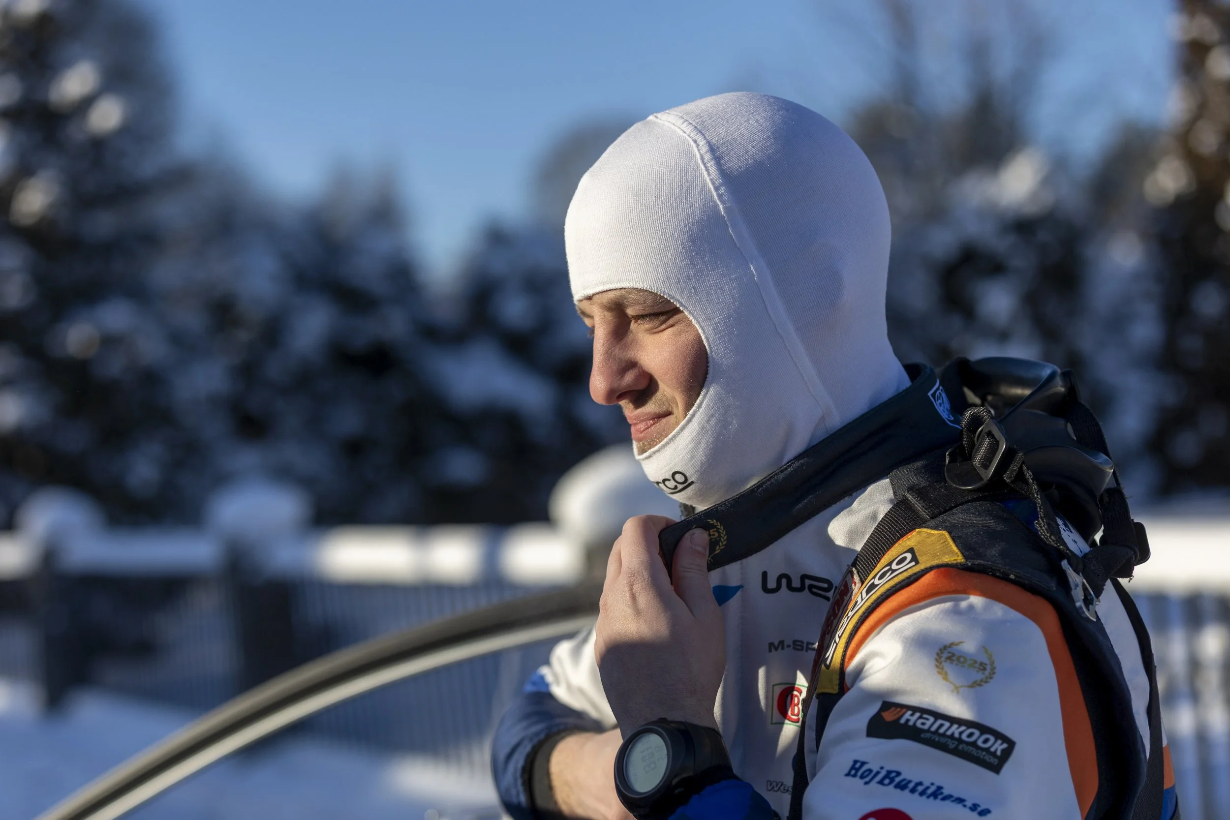 A person dressed in winter sports gear with a white hood, orange, white, and black racing suit, is outdoors in a snowy landscape, possibly preparing for or participating in a winter sports event.