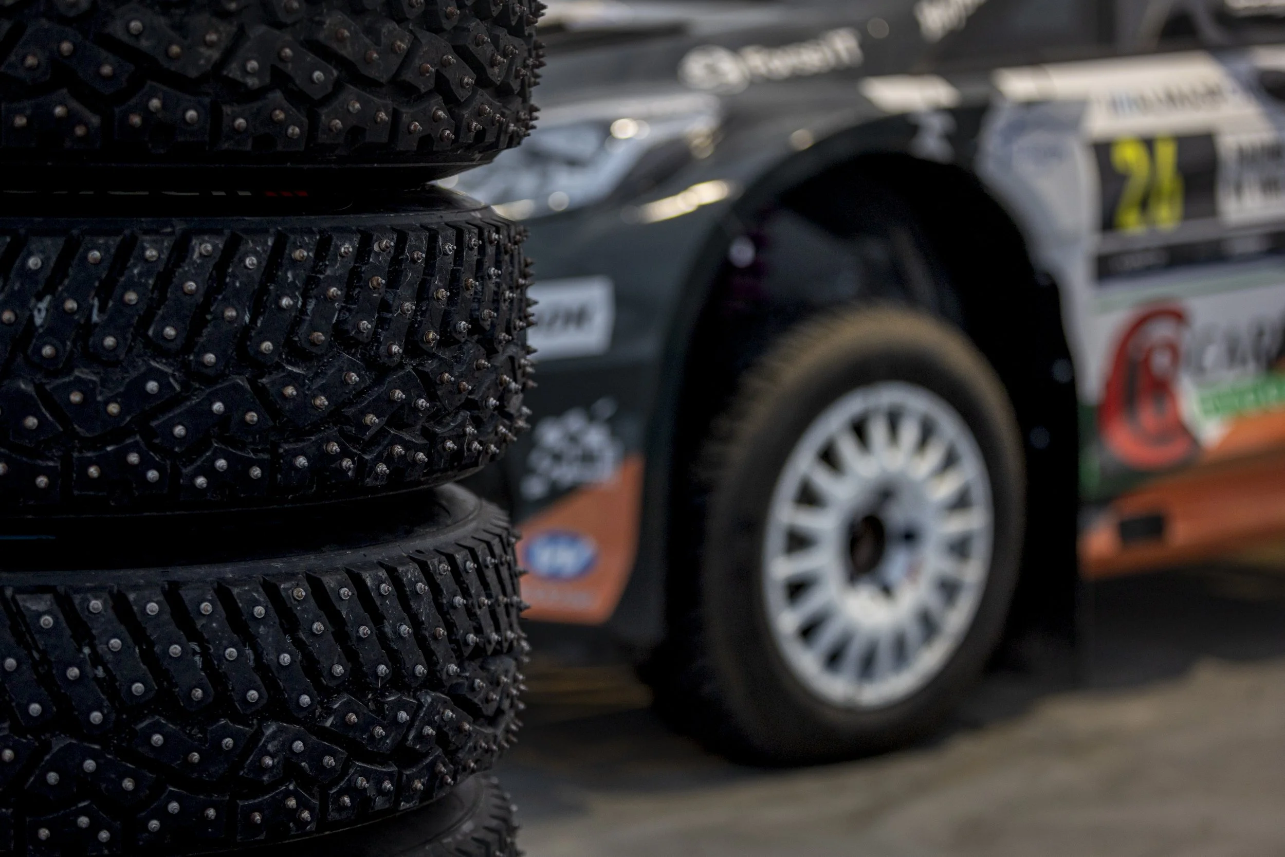 Stacked off-road tires with aggressive tread pattern in the foreground, blurred race cars with sponsor decals in the background.