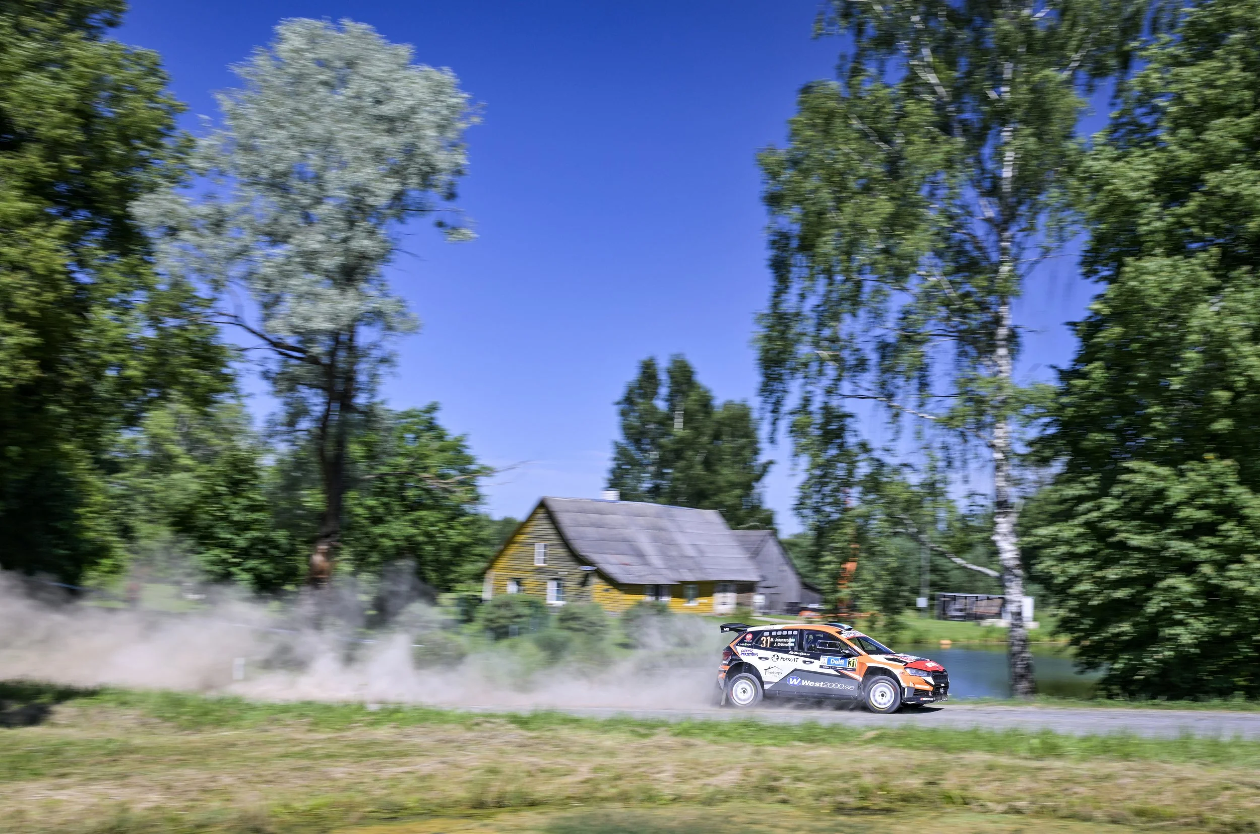 A rally car speeding on a dirt road through a lush green forest with tall trees and a yellow house in the background during daytime.