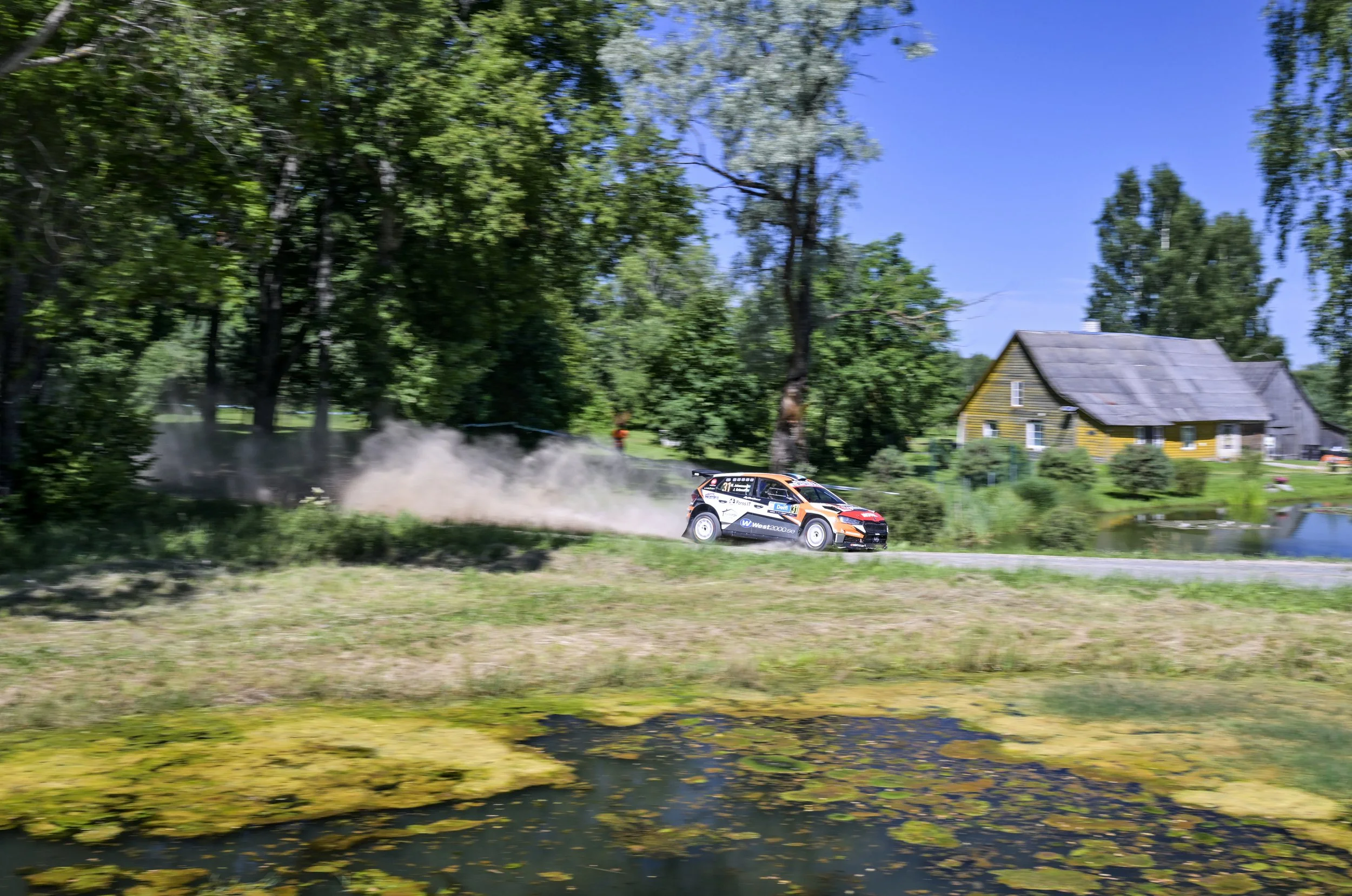 A rally car racing on a gravel road through a green, wooded area with a house and trees in the background, kicking up dust behind it.