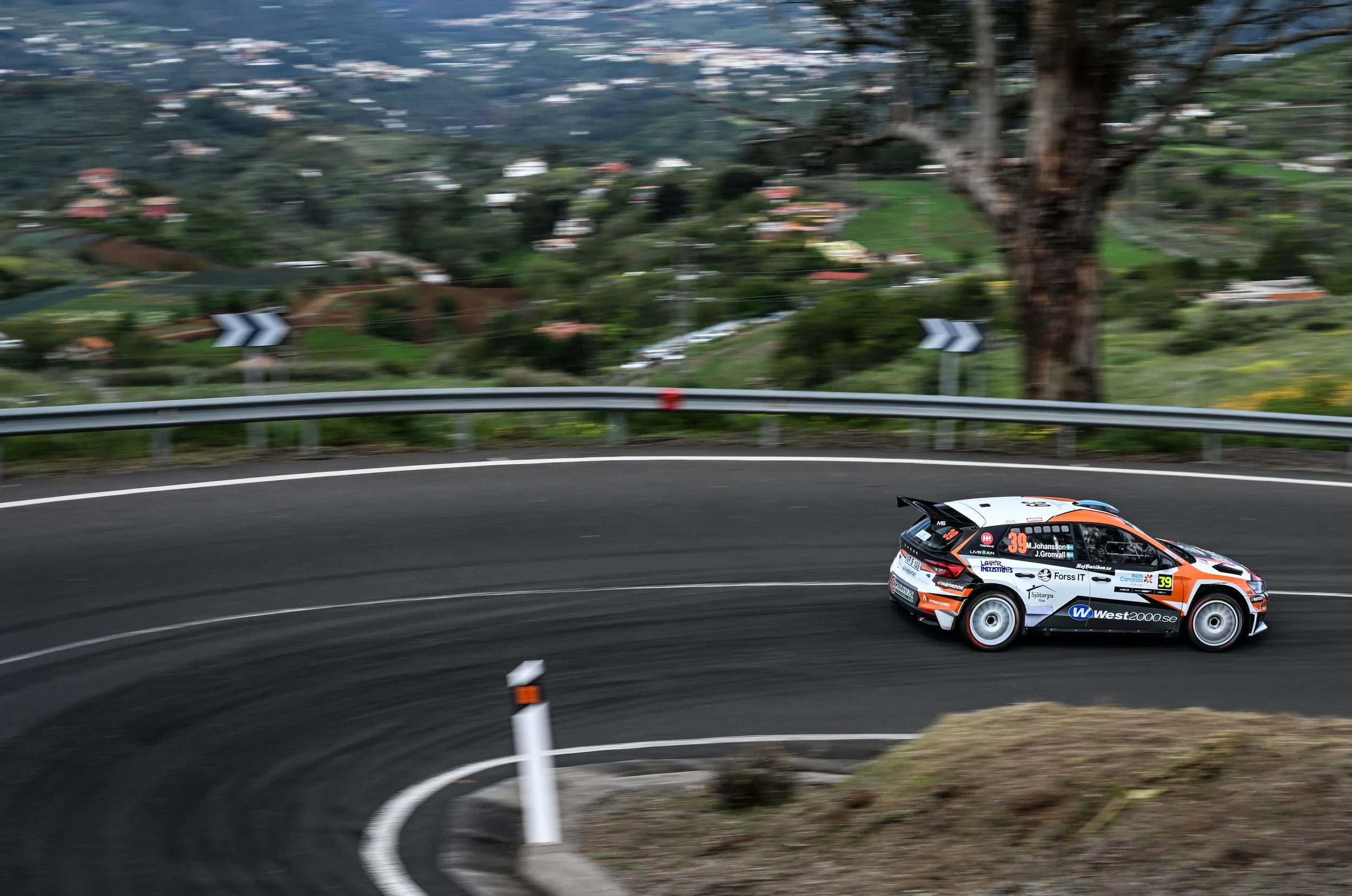 A rally car racing on a curved mountain road with a guardrail, trees, and a blurred suburban landscape in the background.