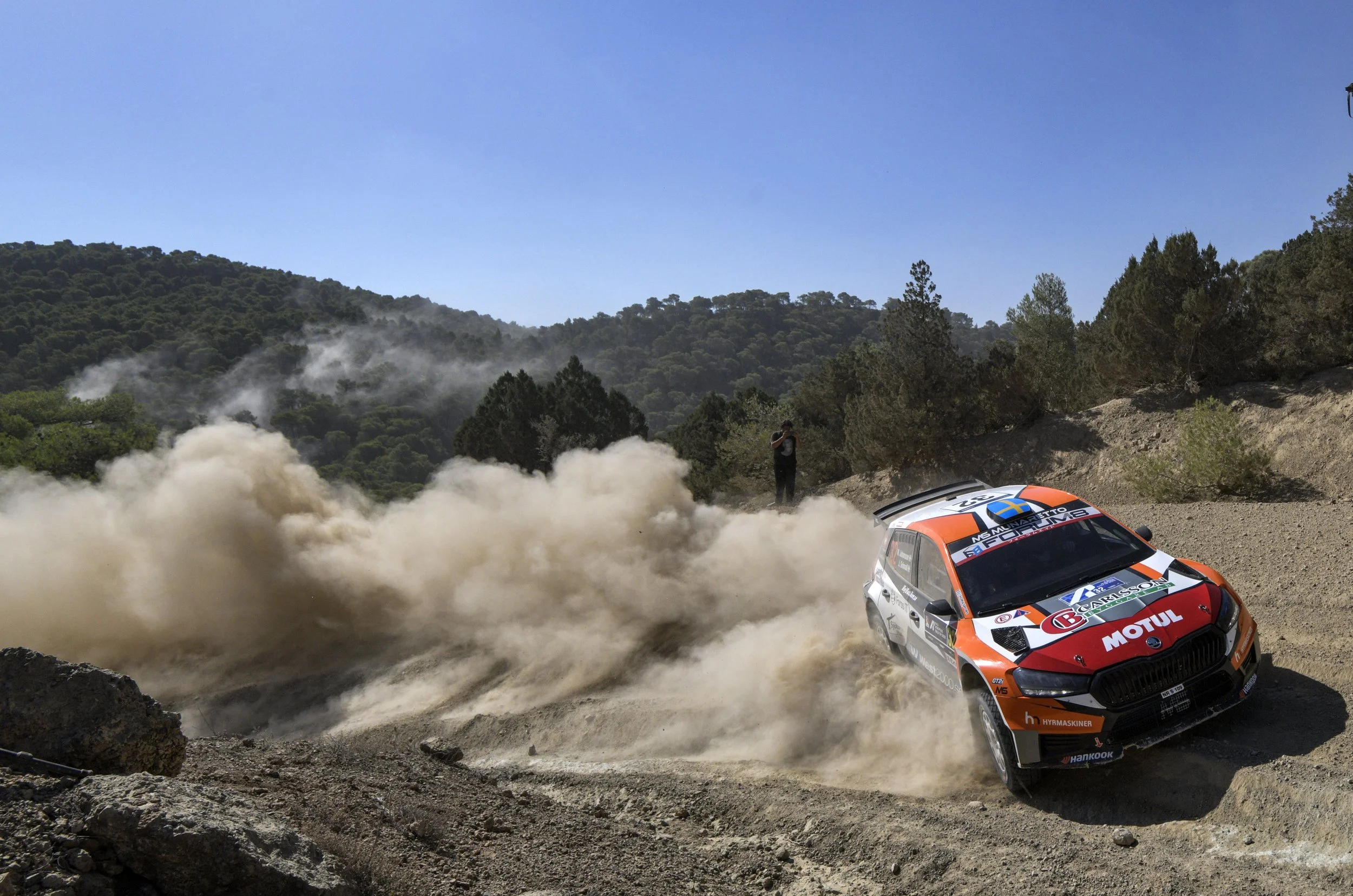 A rally car racing on a dirt road, kicking up dust with a mountainous landscape and a blue sky in the background.