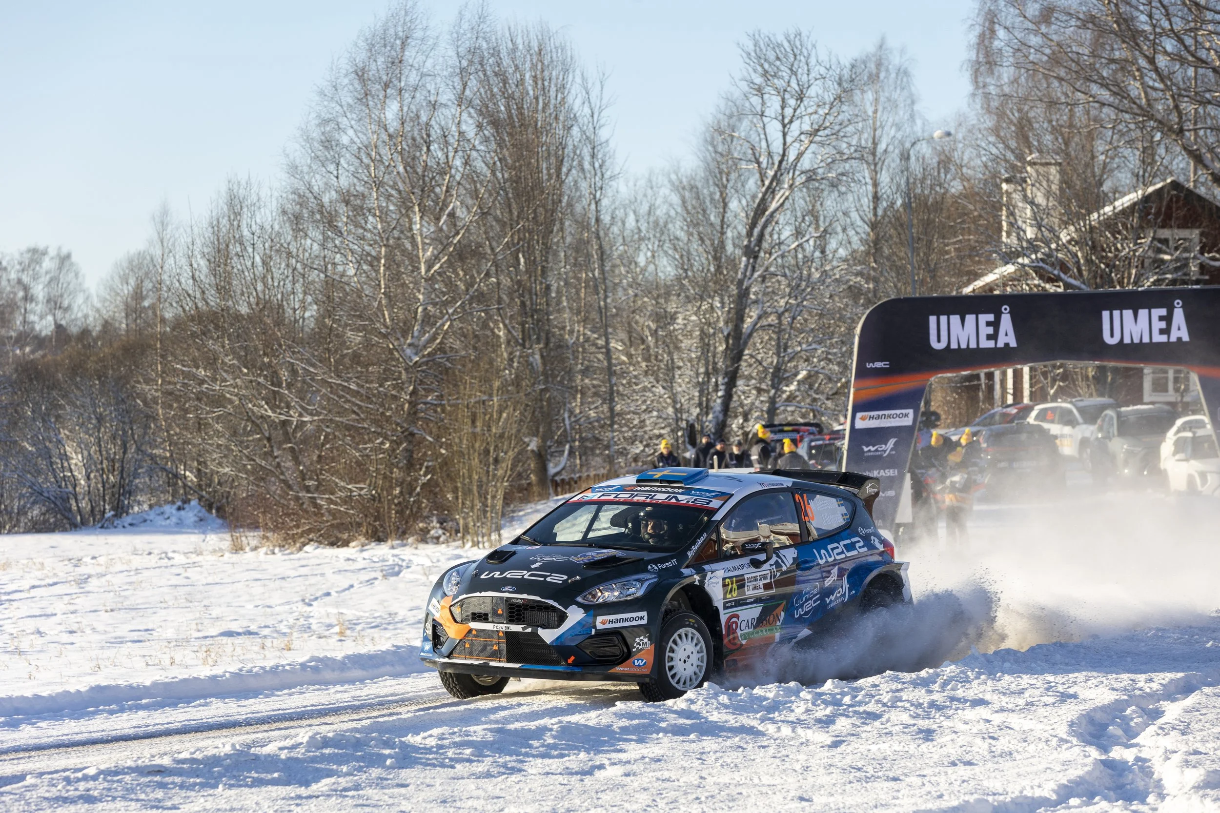 A rally car racing on a snowy track with snow kicking up behind it. The car is black and blue with various sponsor decals. In the background, there is a snowy landscape with trees and a rally stage arch that reads 'UMEA'.