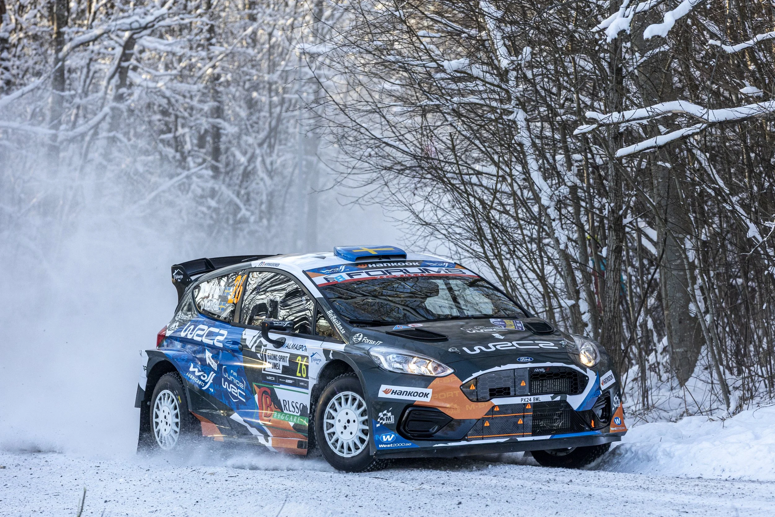 A rally car driving on a snowy forest road during a winter race, creating a spray of snow behind it.