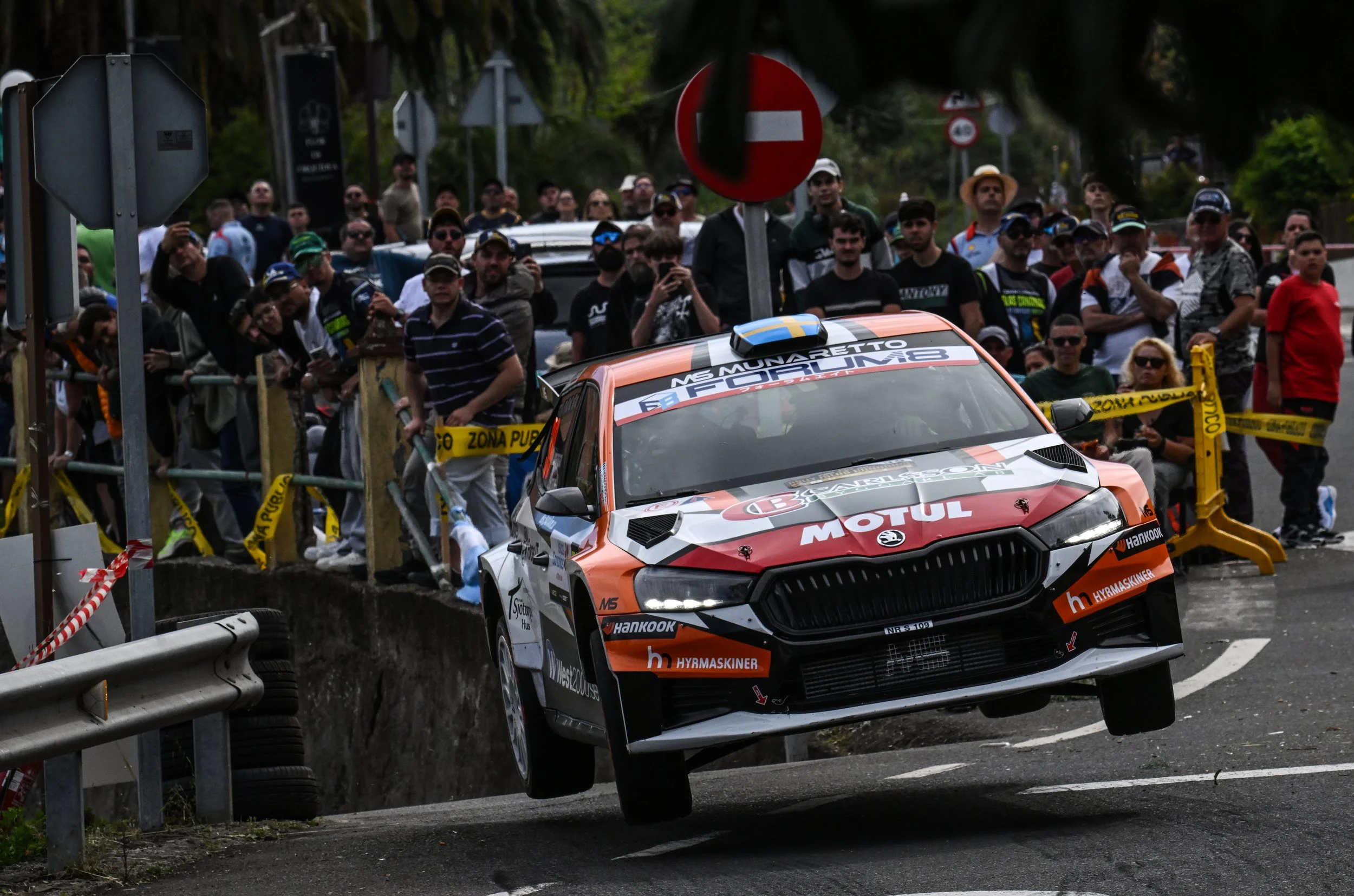 A rally car is jumping over a bump on a road surrounded by a crowd of spectators behind a barrier, with warning signs and trees in the background.