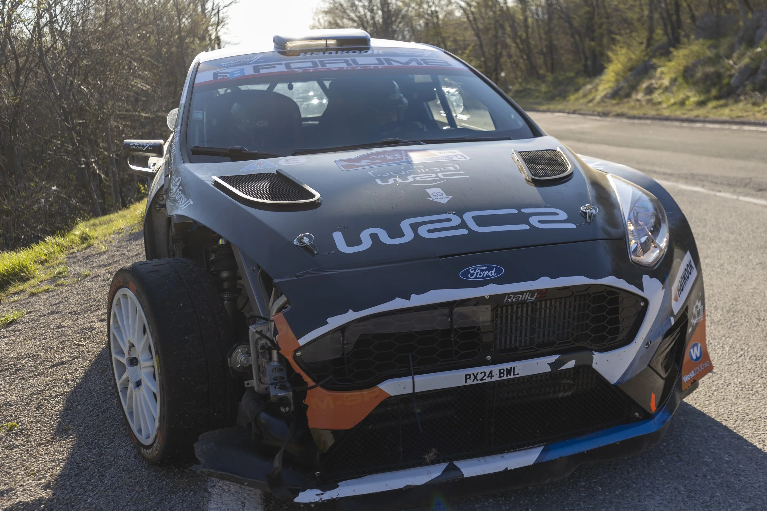 A rally car on a dirt and paved roadside with trees in the background. The car features sponsor logos including Ford and wetsc. It is in motion, with its front wheels turned sharply.