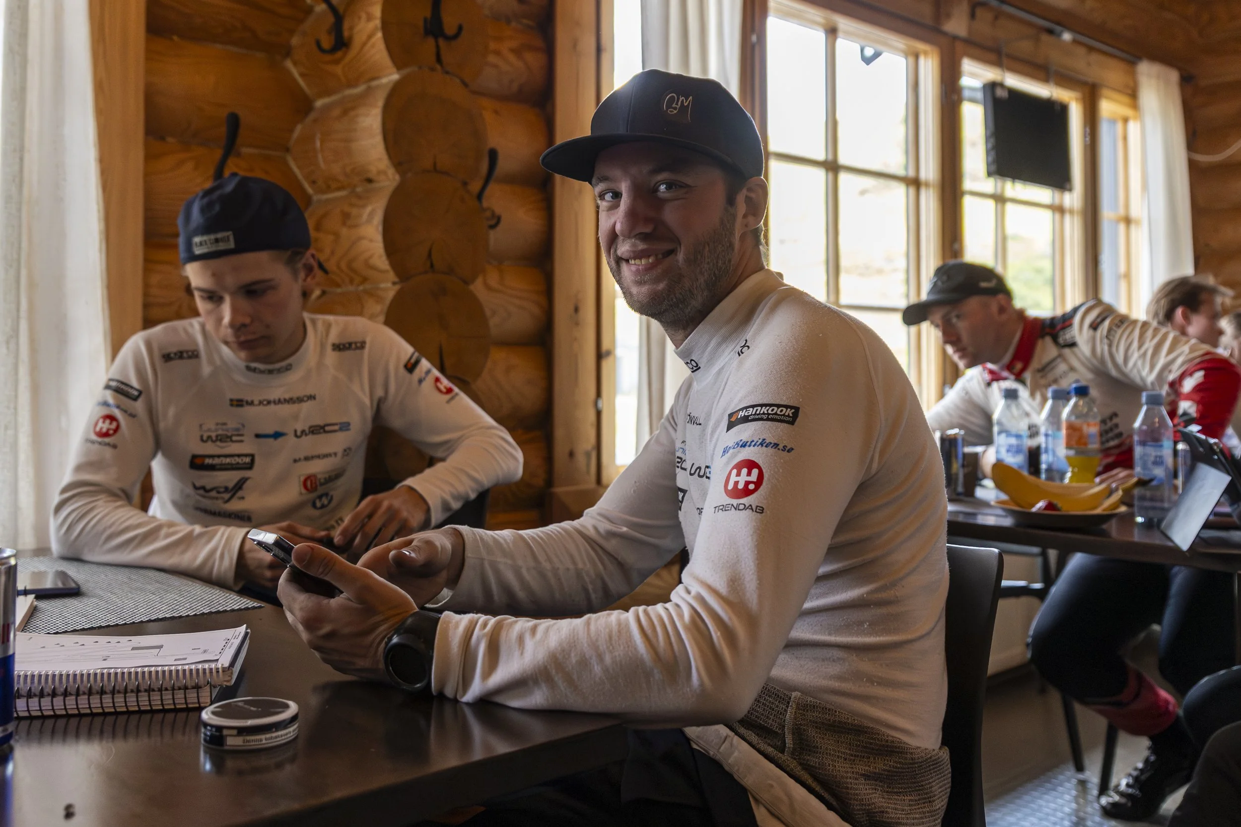 A man in a black cap smiling at the camera while sitting at a table with a racing team in a wooden cabin-style room. Other team members are focused on their tasks, with tools and water bottles on the table. The room has large windows and wood panelin