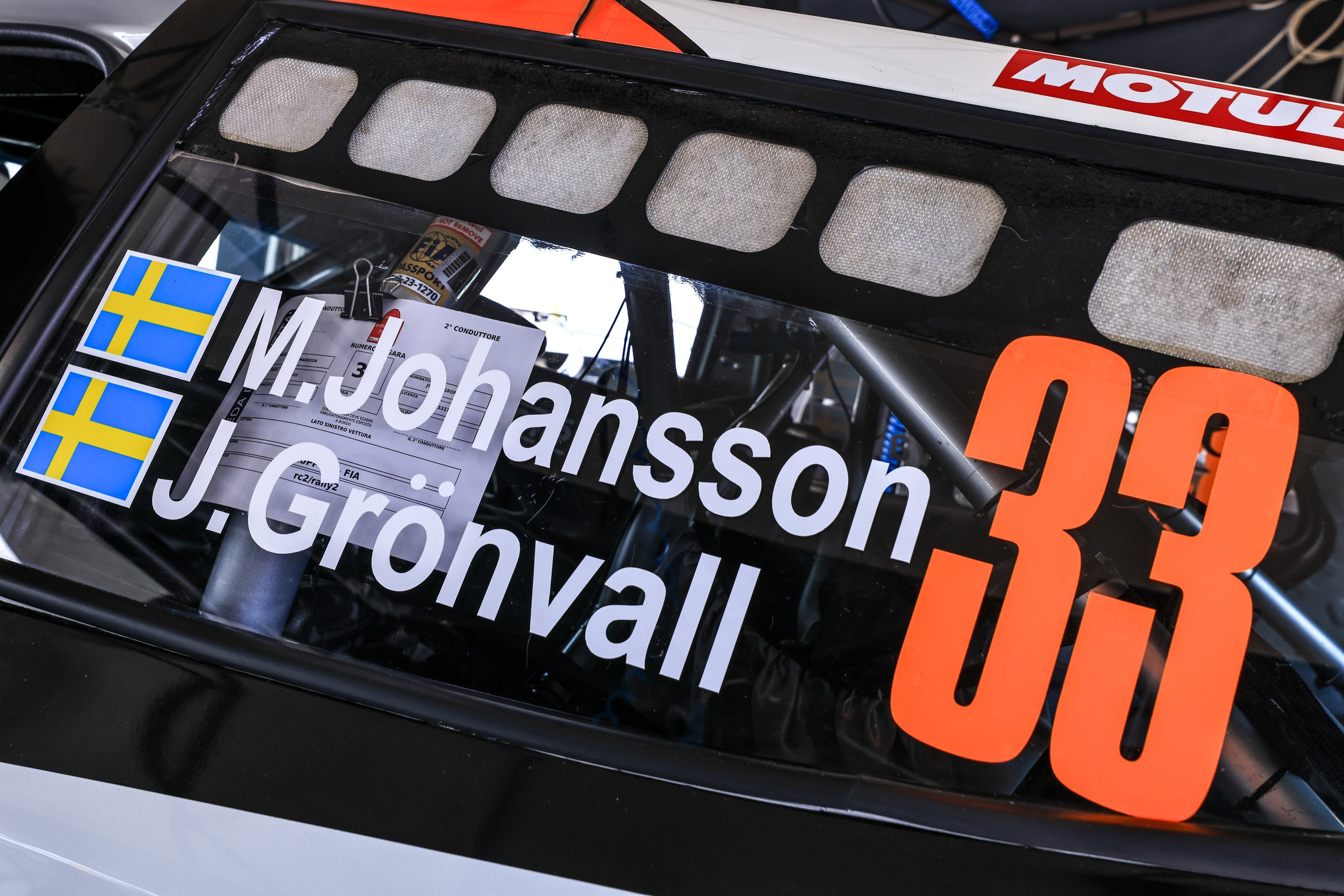 Close-up image of a race car windshield with multiple decals, including Swedish flags, a large orange number, and sponsor logos such as Motul. The top portion shows a black vent with mesh covering it.