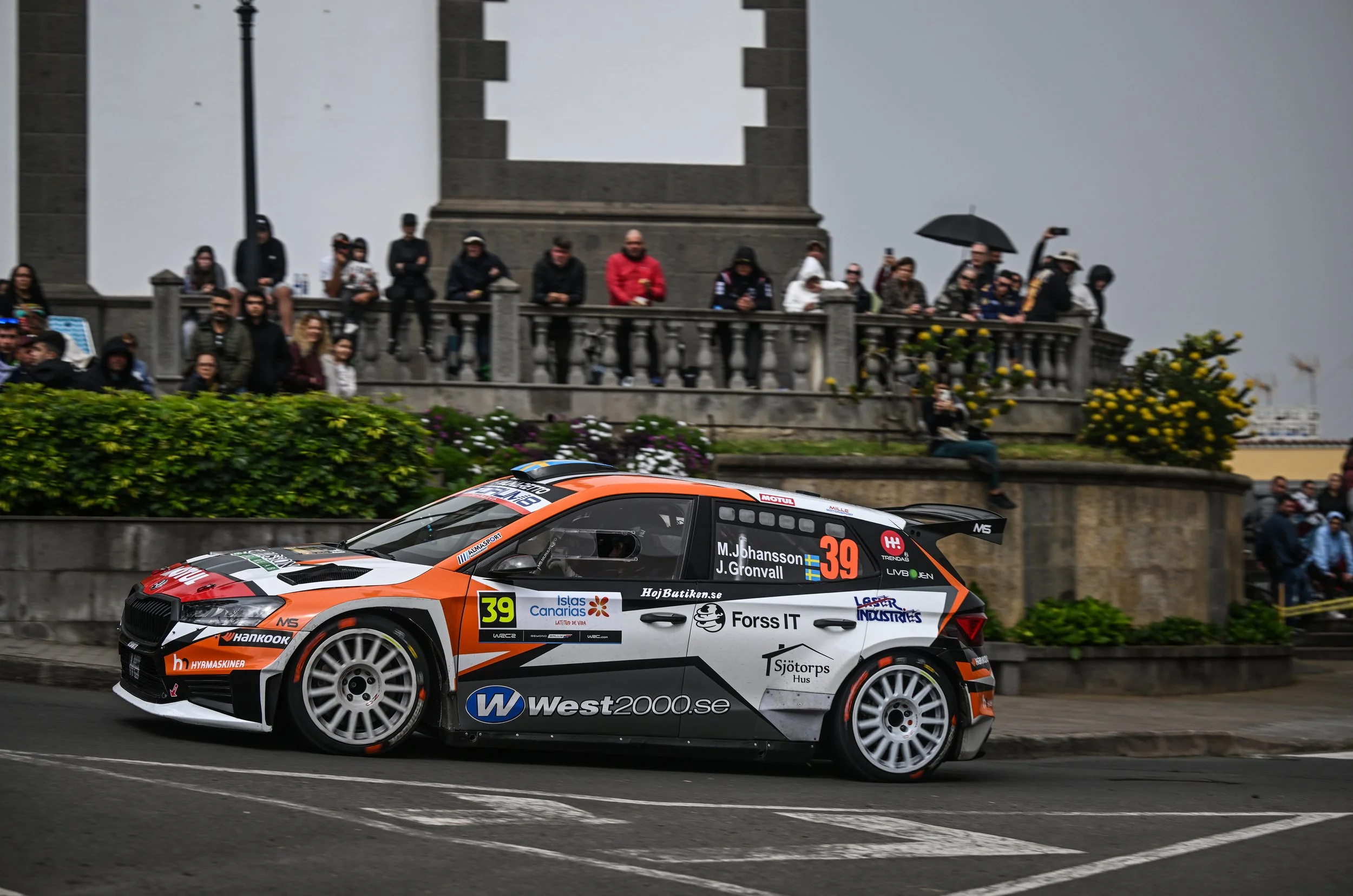 A rally car racing on a city street with spectators watching from a balcony and sidewalk. The car is orange, black, and white.
