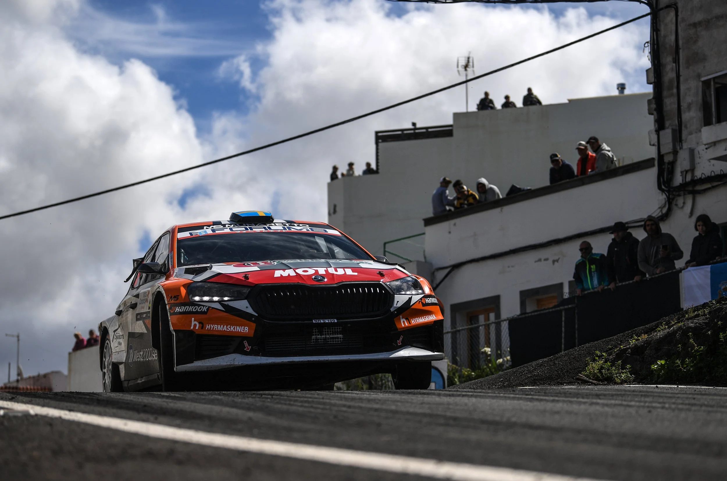 Orange and black race car with sponsorship logos speeding down a road, with spectators watching from buildings above.