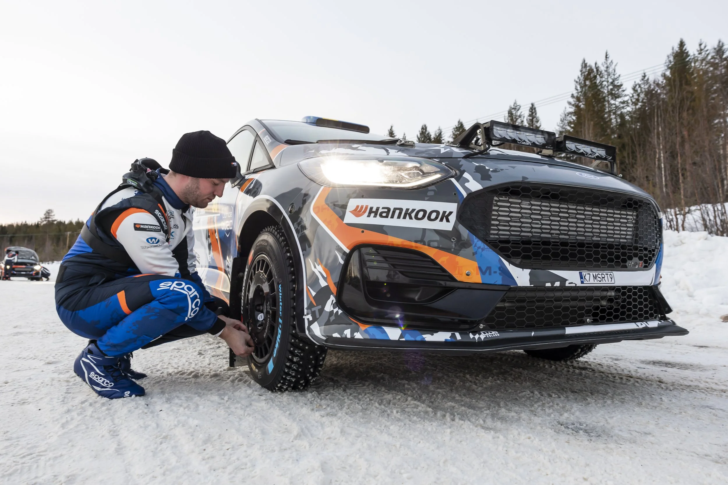 A rally driver adjusting the wheel of a Rally car on snow with a forest in the background.