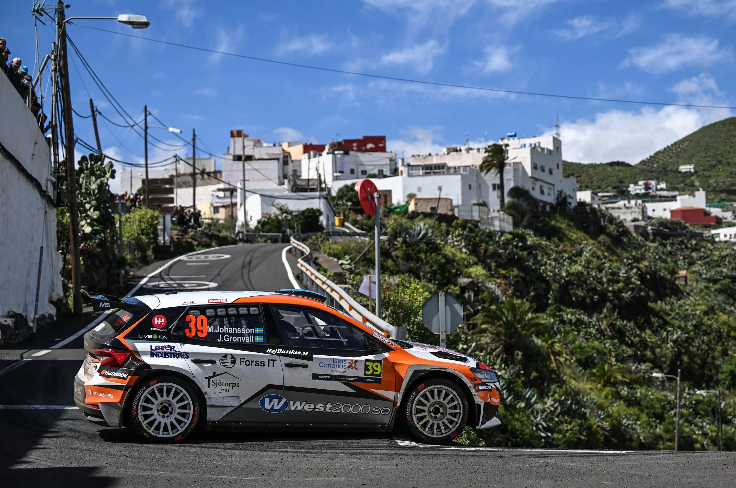 A rally car with race number 39 driven by M. Johansson and J. Gronvall is racing on a narrow, winding road in a hilly area with white buildings and lush green vegetation in the background. The sky is blue with some scattered clouds.