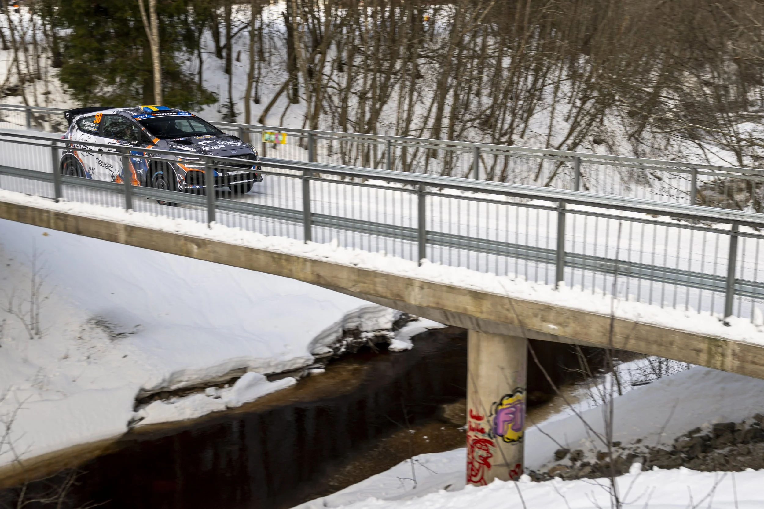 A race car driving on a snow-covered road next to a forest with leafless trees and a small stream underneath a bridge, with graffiti on a post near the stream.