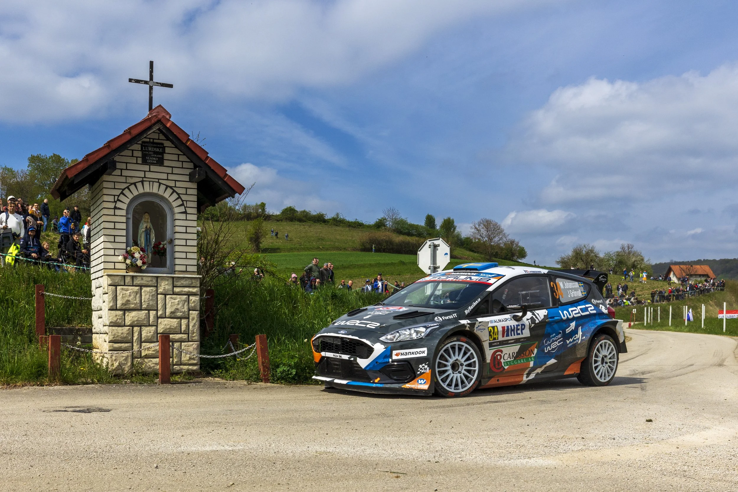 A rally car speeding on a dirt road with a crowd of spectators and a small shrine with a statue of the Virgin Mary nearby, against a background of green hills and a partly cloudy sky.