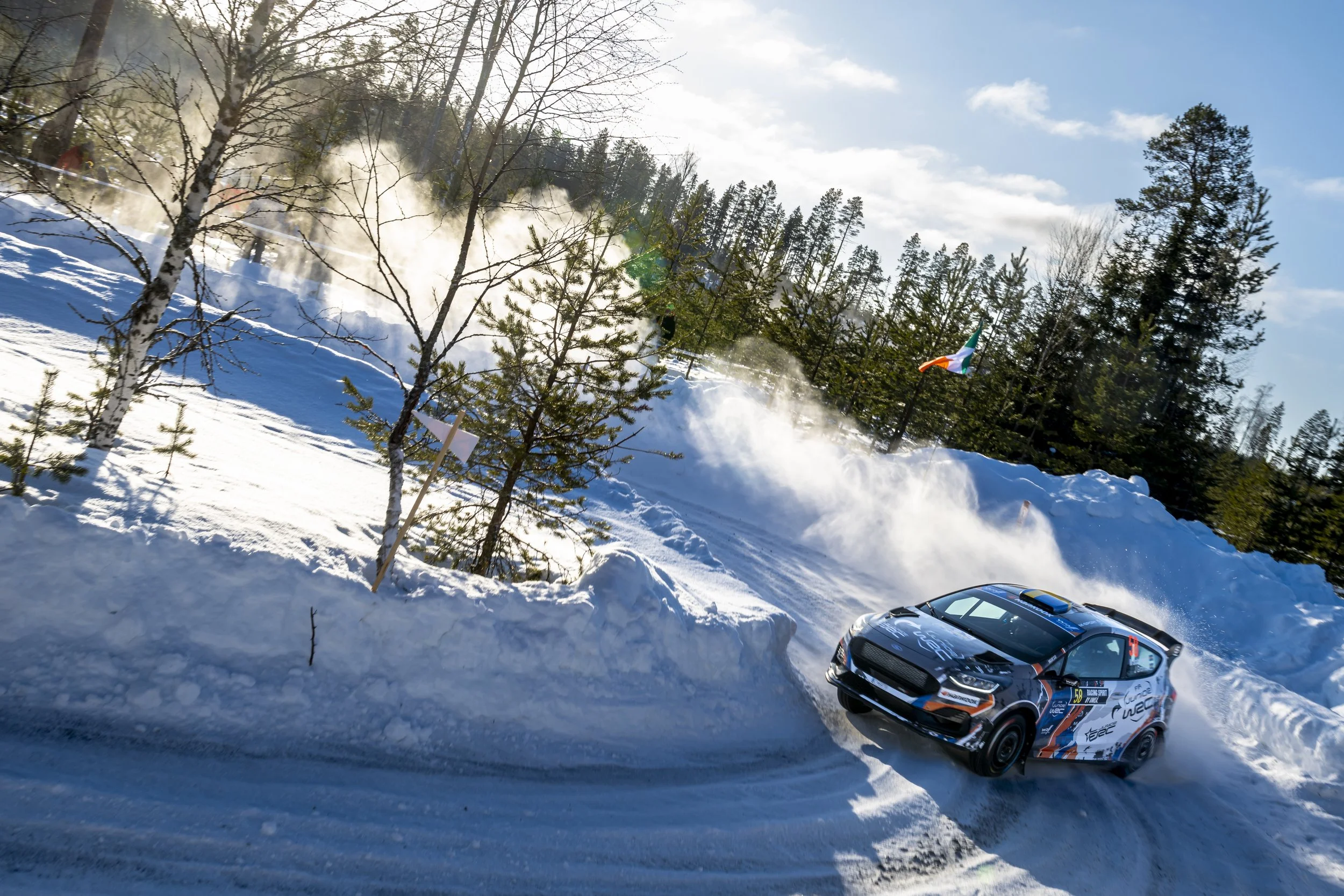 A rally car racing on a snowy mountain road with trees and a blue sky in the background, emitting snow and dust as it speeds.
