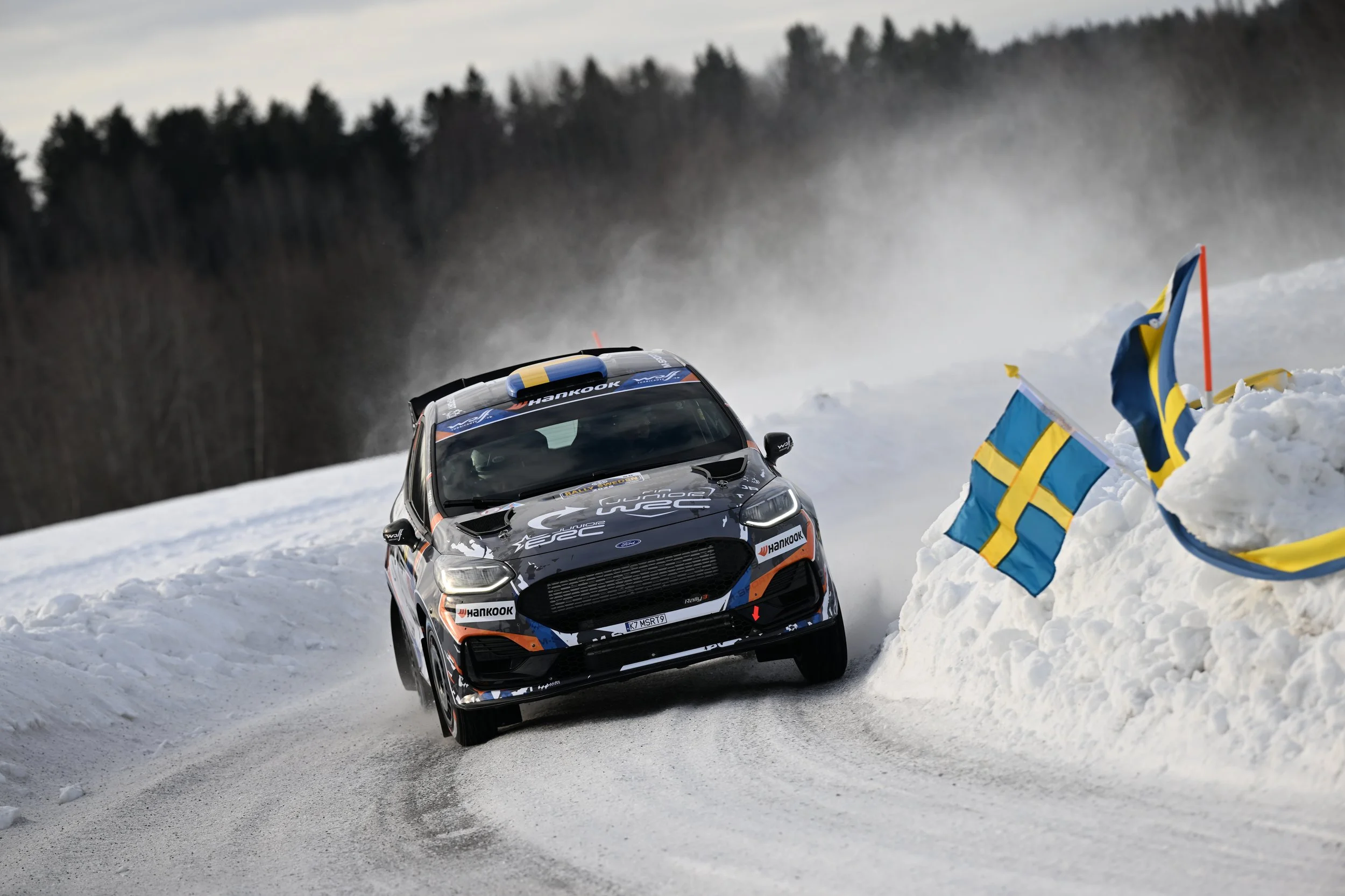 A race car speeding on an icy snow-covered track, with Swedish flags on the side and snow banks on the edges.