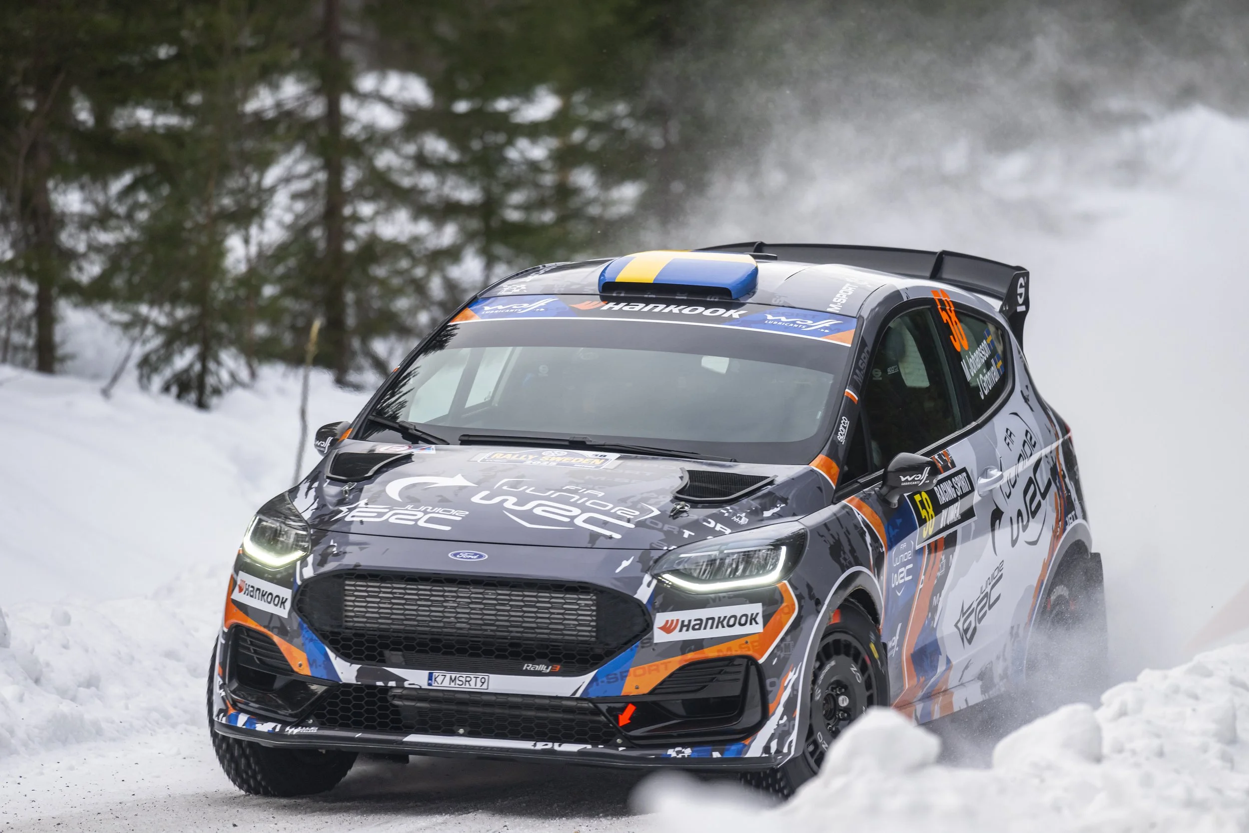 A black rally car with blue, orange, and white graphics racing on snowy terrain, surrounded by snow-covered ground and pine trees.