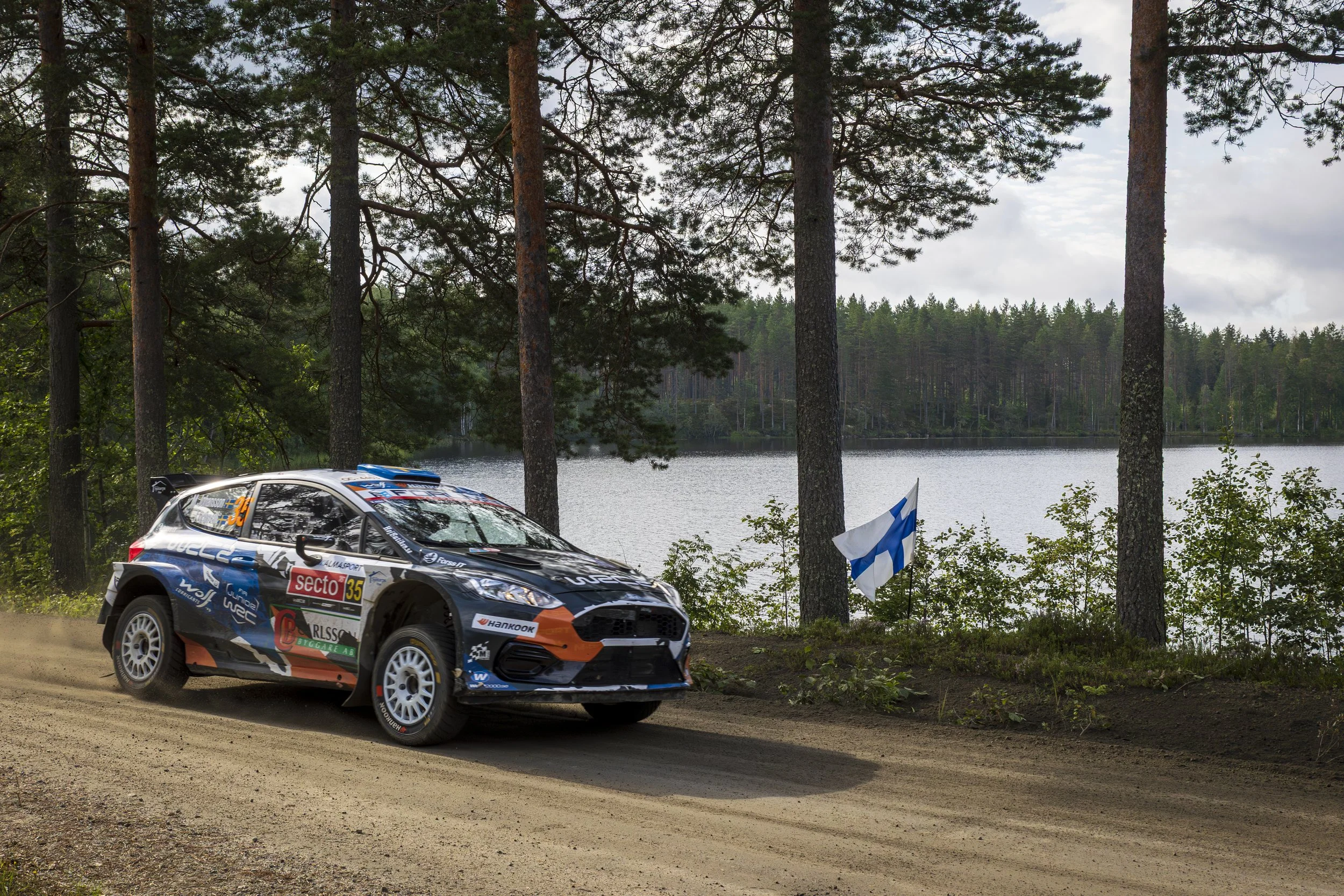 A rally car with racing decals and the number 35 is driving on a dirt road beside a lake, with a forest of tall pine trees in the background and Finnish flags hanging from a bush.