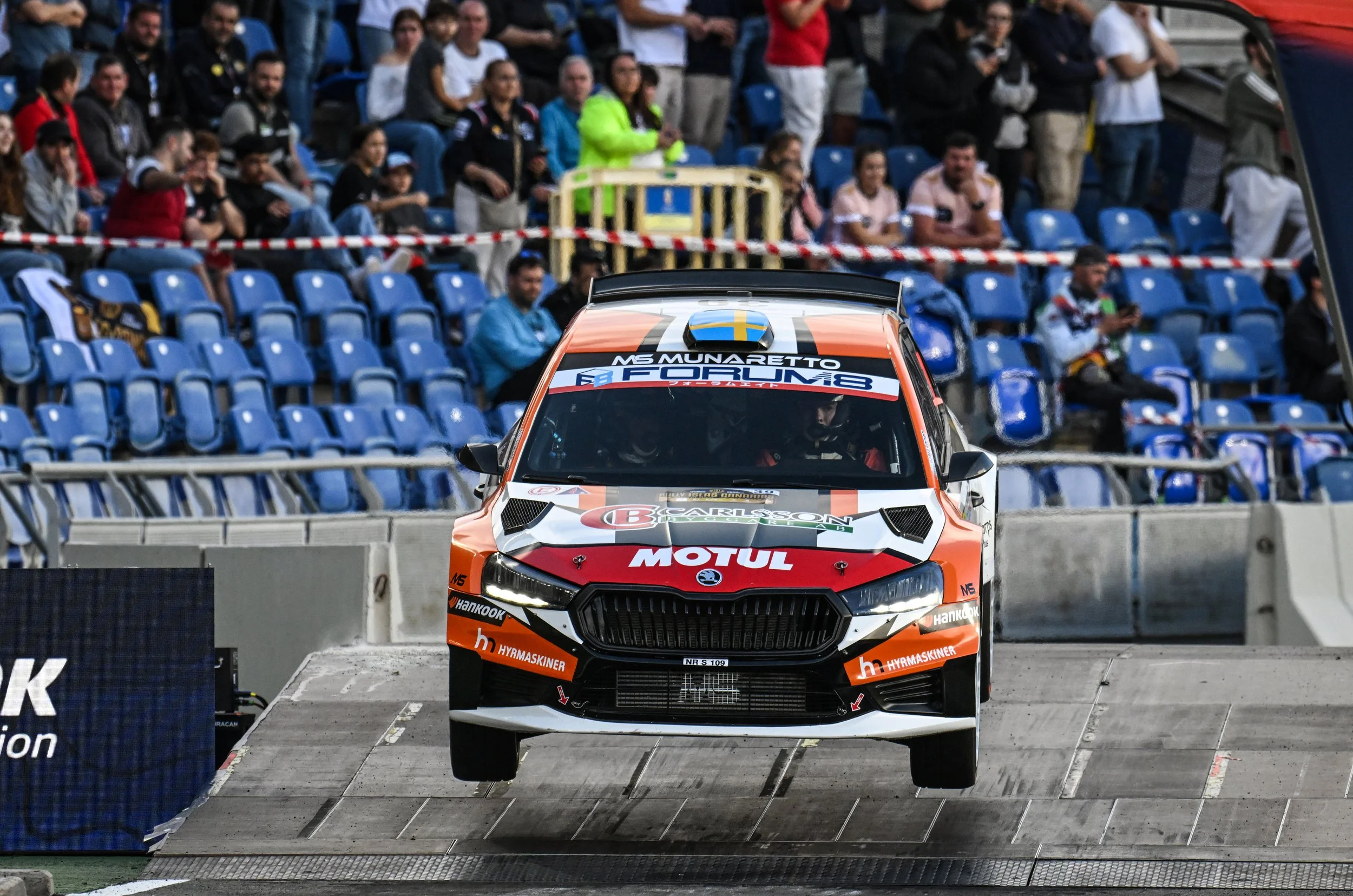 A rally car with sponsor logos and orange and black livery launching off a ramp during a racing event, with spectators in the background.